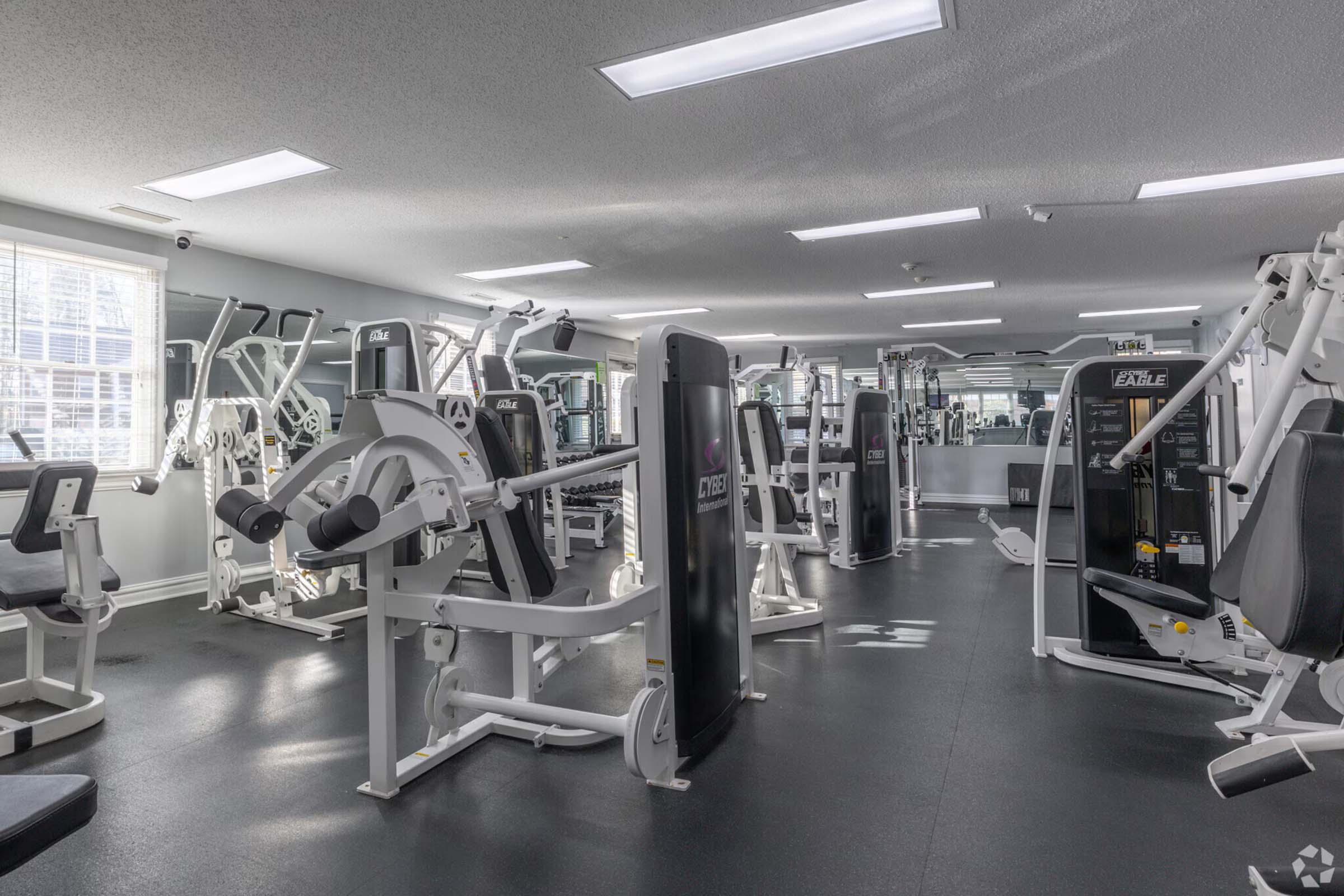 A well-lit gym filled with various workout equipment, including weight machines, benches, and exercise machines. The floor is covered with dark rubber matting, and there are mirrors on the walls reflecting the equipment. Large windows allow natural light to enter the space.