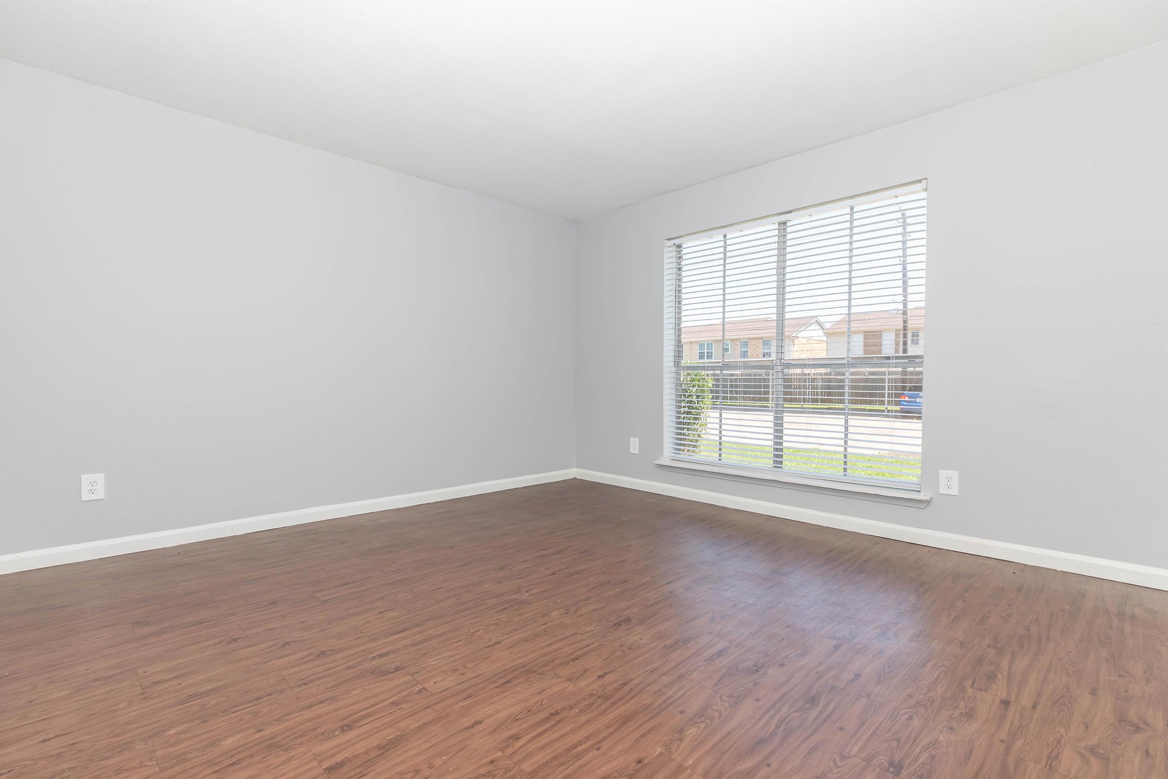 A spacious, empty room with light gray walls and a large window featuring white blinds. The floor is finished with a wood-like laminate, creating a warm atmosphere. There are no furnishings, highlighting the open space and natural light coming in through the window.