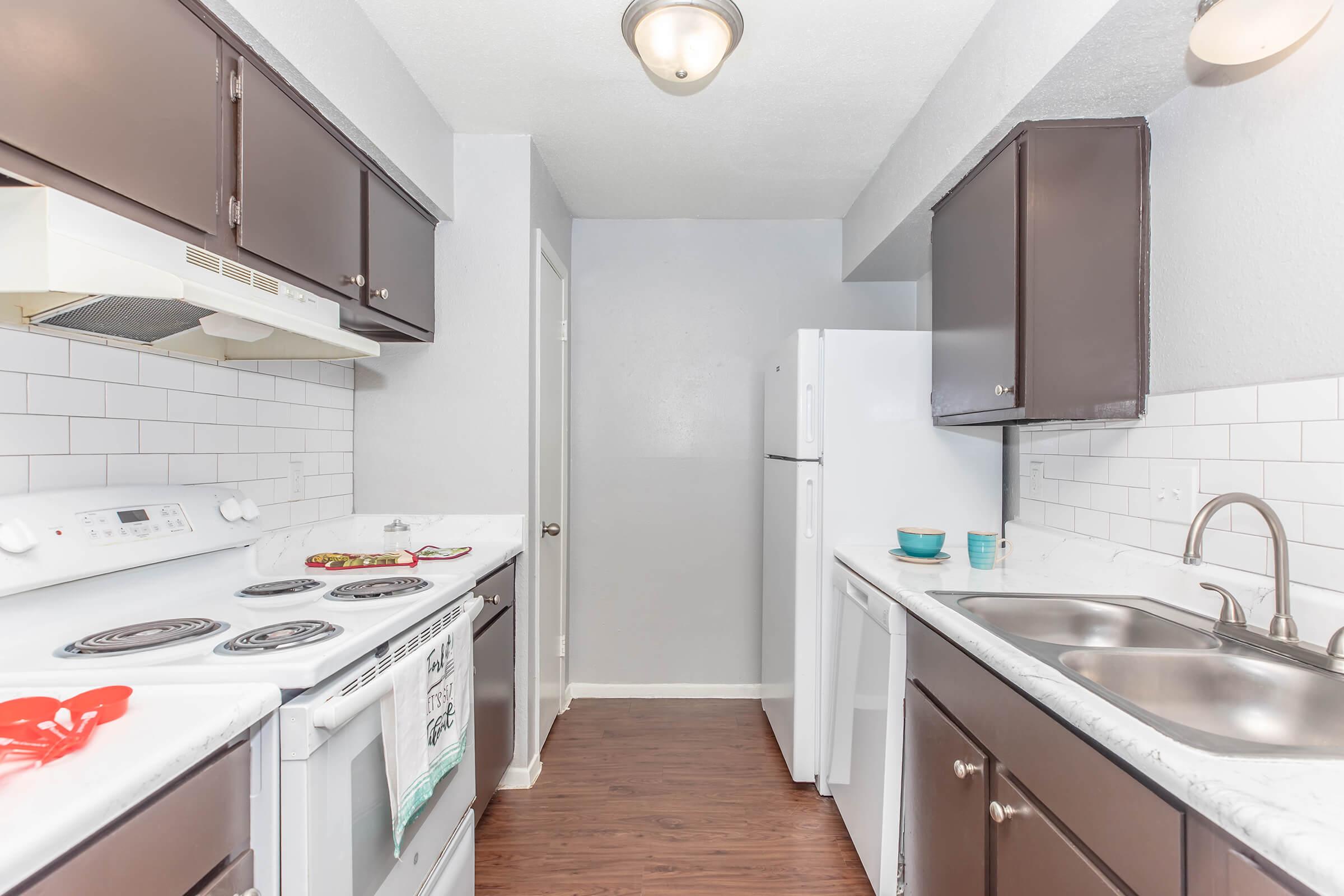 A small, modern kitchen featuring dark cabinetry, a white stove and refrigerator, and a double sink. Light gray walls and white backsplash tiles create a clean, bright atmosphere. A door leads to another room, while a few items are placed on the countertop, adding a touch of homey decor.