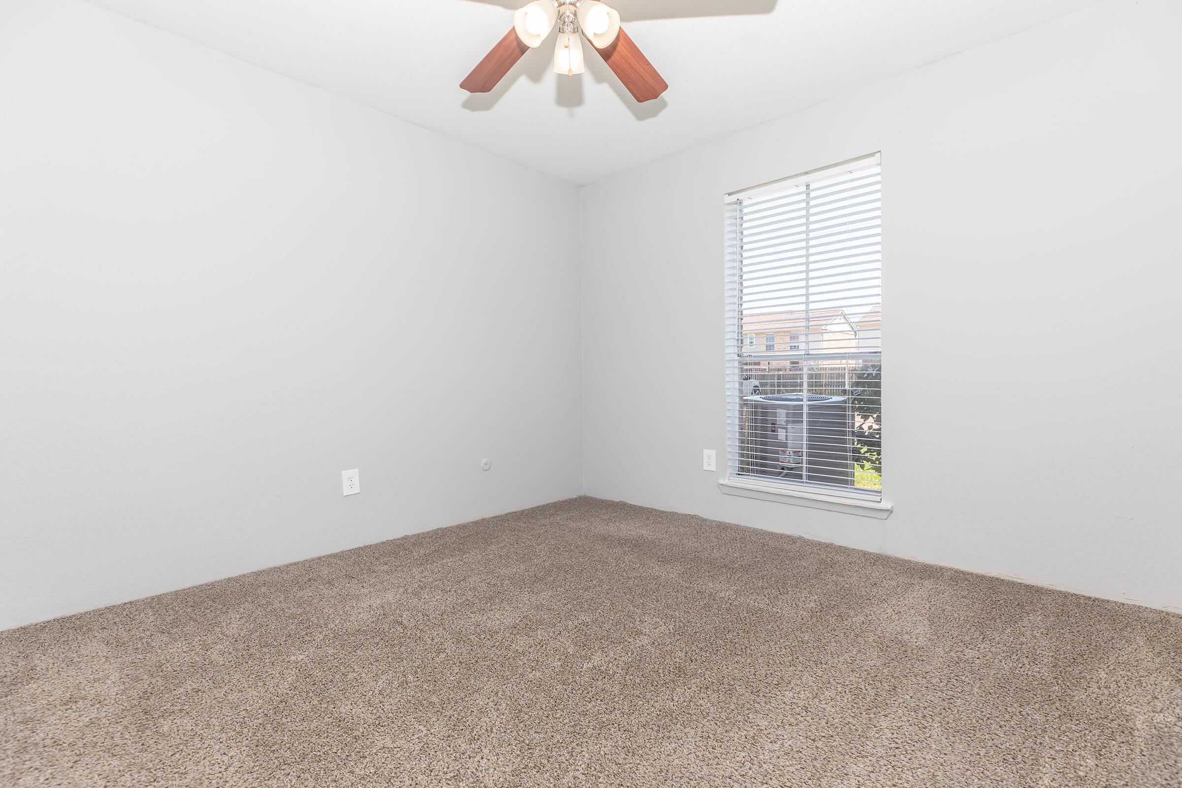 A vacant room with light gray walls and a ceiling fan, featuring a window covered with blinds. The floor is carpeted in a soft texture, and there are no furnishings or decorations present, creating a spacious and airy ambiance.