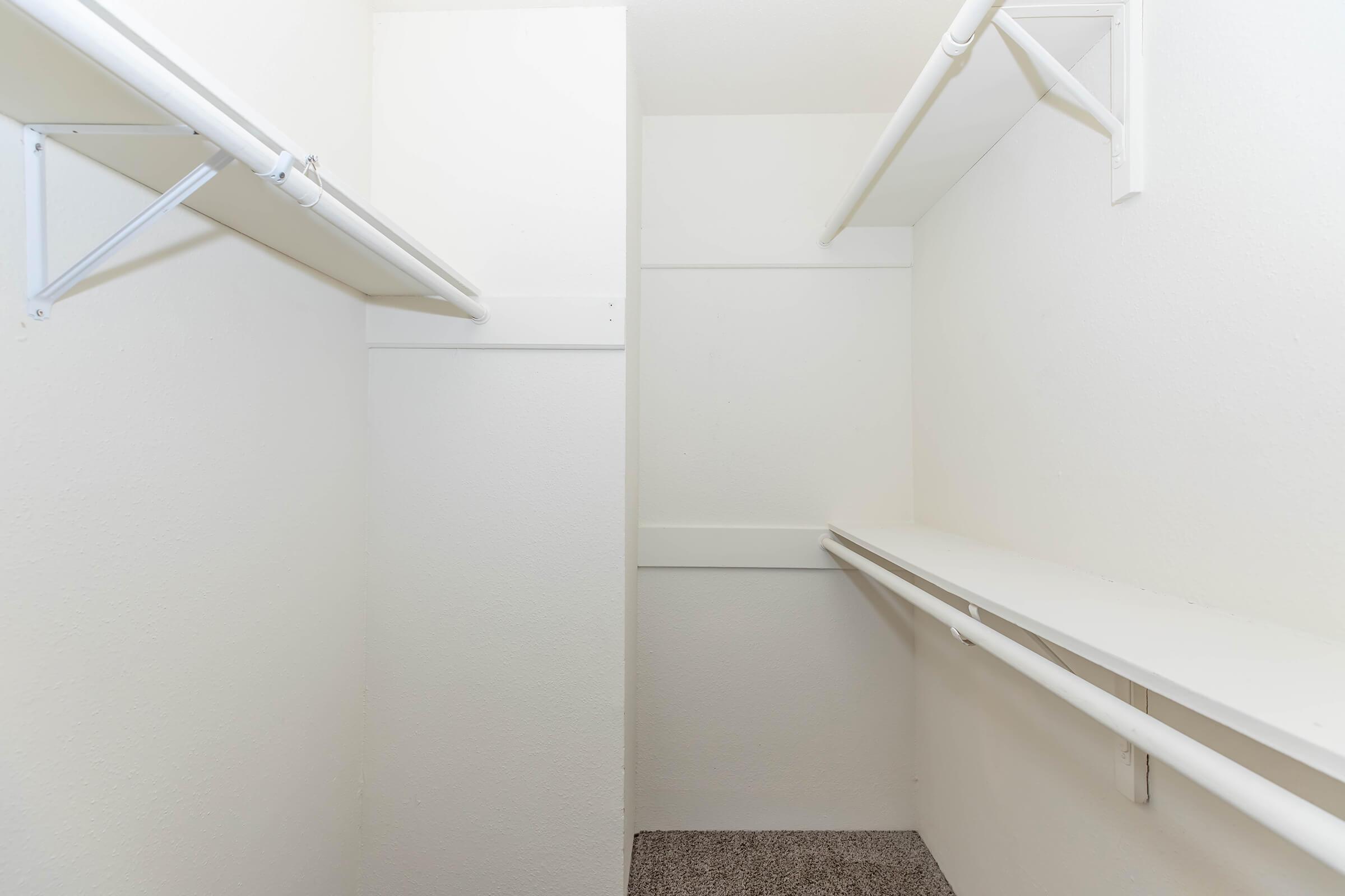 Empty closet with white walls and a grey carpet. There are two hanging rods on opposite walls and a shelf along one side, providing ample storage space. The overall appearance is clean and tidy, with a minimalist design.