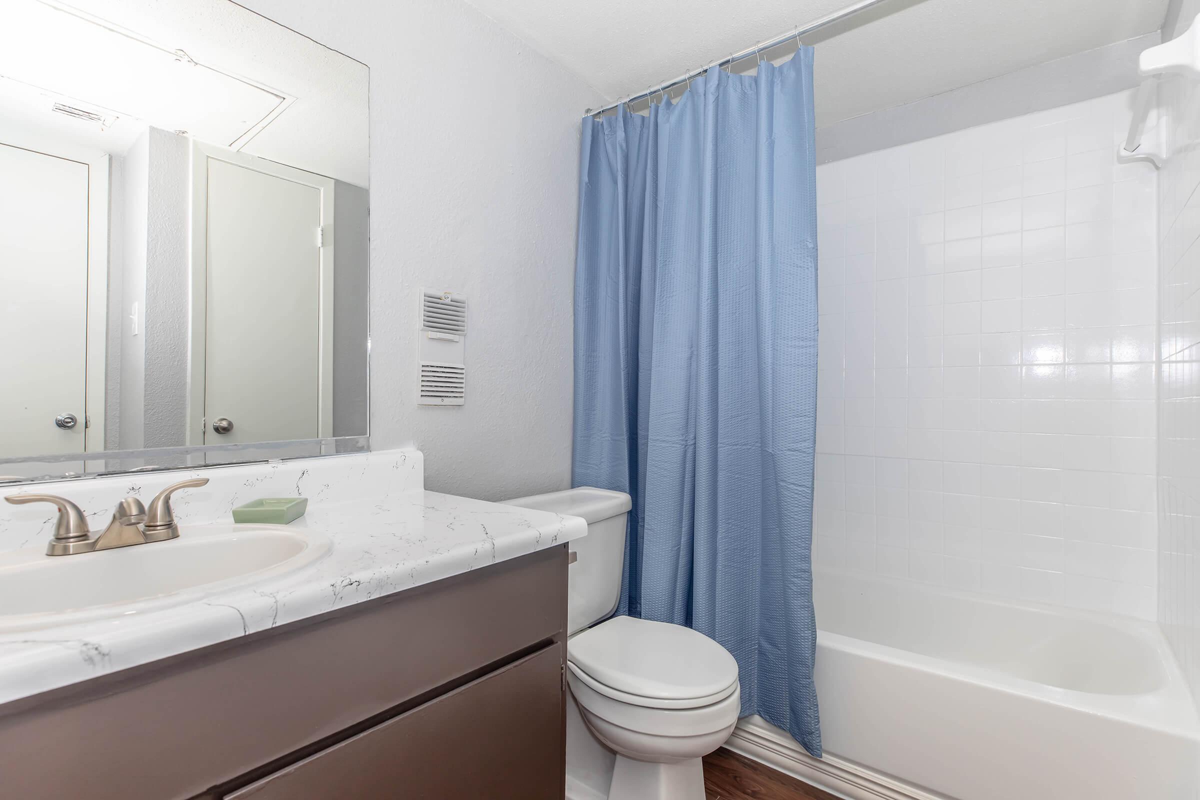 A clean, modern bathroom featuring a bathtub with a shower, a toilet, and a sink with a marble countertop. The walls are painted light gray, and a blue shower curtain adds a pop of color. A mirror above the sink and light-colored flooring complete the space.