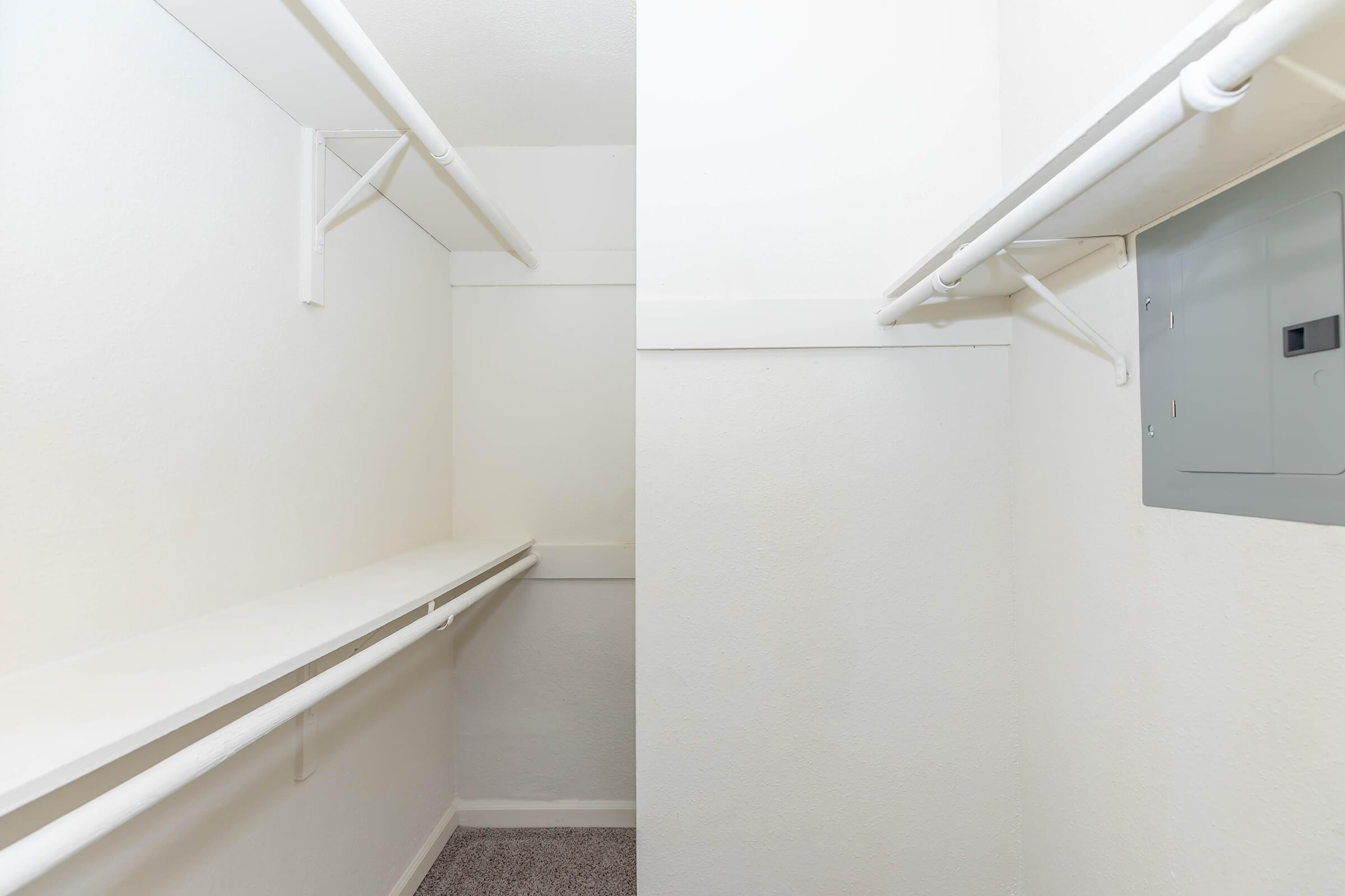 Empty closet space with white walls, a long shelf along one side, and a gray electrical panel mounted on the wall. The floor is carpeted, and the overall lighting is bright and neutral, creating a clean and minimalist appearance.