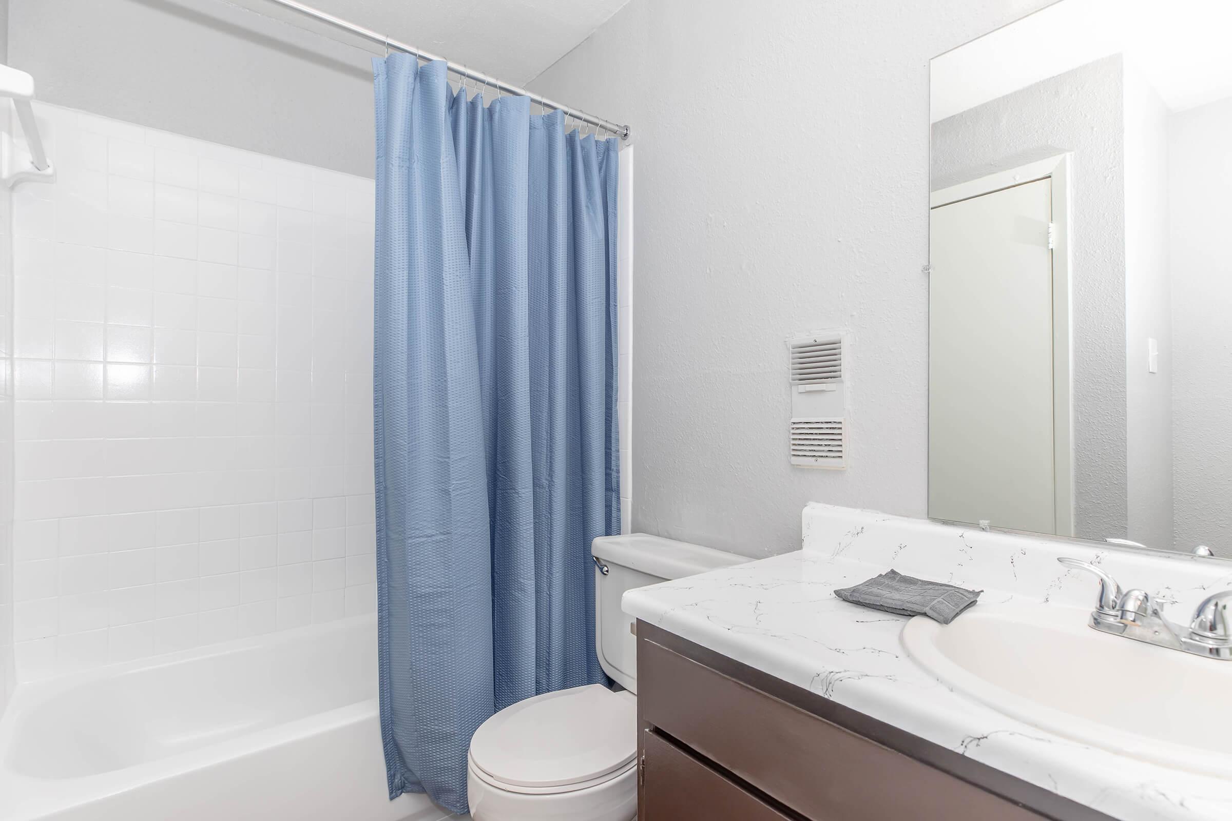 A clean and simple bathroom featuring a white bathtub with a blue shower curtain, a white toilet, and a sink with a marble countertop. The walls are painted in a light gray color, and there is a mirror above the sink. A gray hand towel is placed on the counter, and a bathroom vent is visible on the wall.