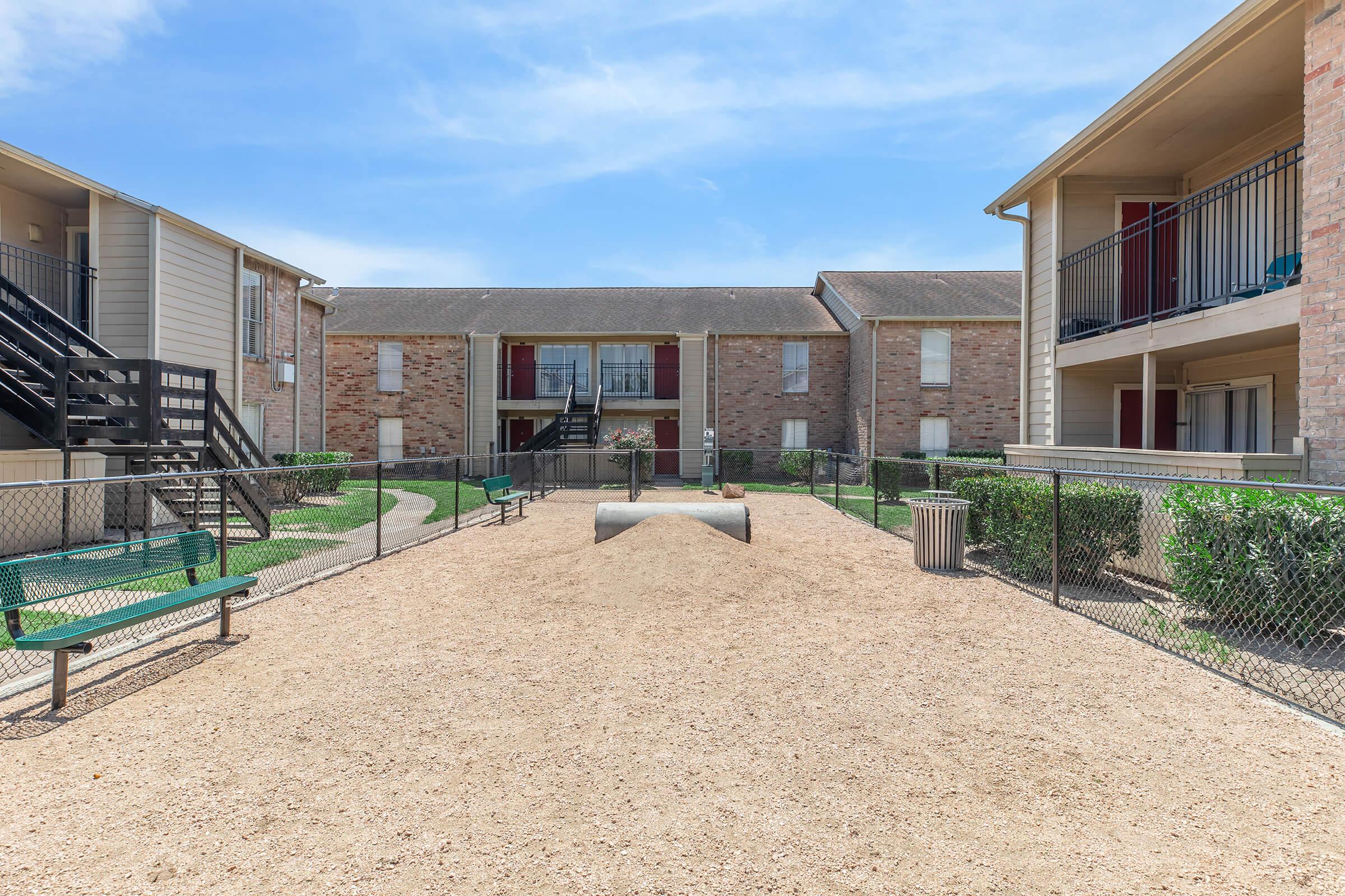 View of a small outdoor courtyard at an apartment complex with two-story buildings on either side. The courtyard features a sandy ground, benches, and a central playground area with a tunnel structure, surrounded by green shrubs and a chain-link fence. Bright blue sky overhead.