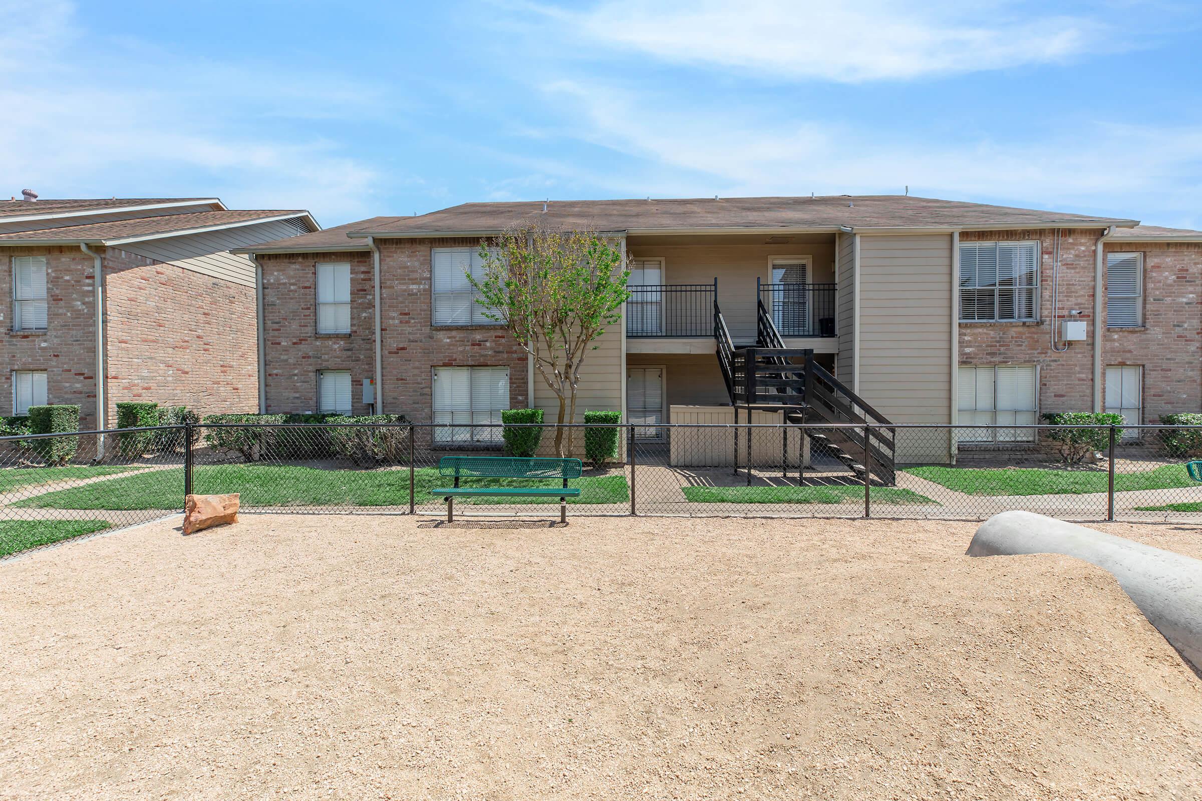 Two multi-unit residential buildings made of brick with a grassy area in front. A wooden staircase leads to the second floor of one building. A small fenced area with sand is visible in the foreground, along with a bench and a small tree. The sky is partly cloudy, indicating a pleasant day.