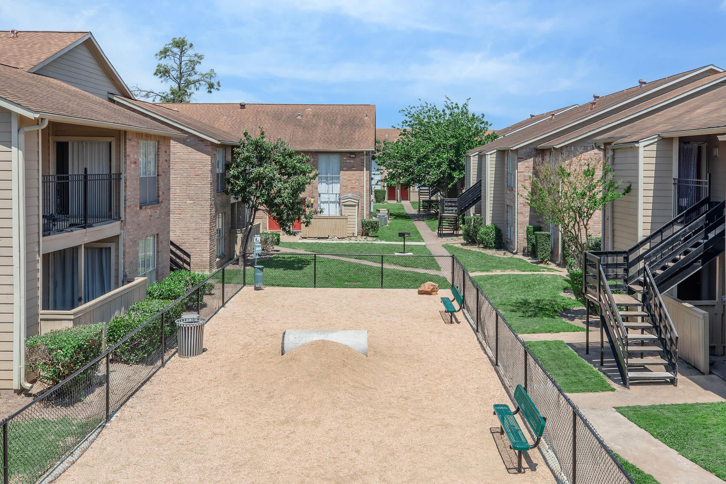 View of an apartment complex courtyard featuring two rows of apartments, a sandy area with a small mound, benches along a pathway, and green grass. Trees provide shade, and the sky is clear with a few clouds.