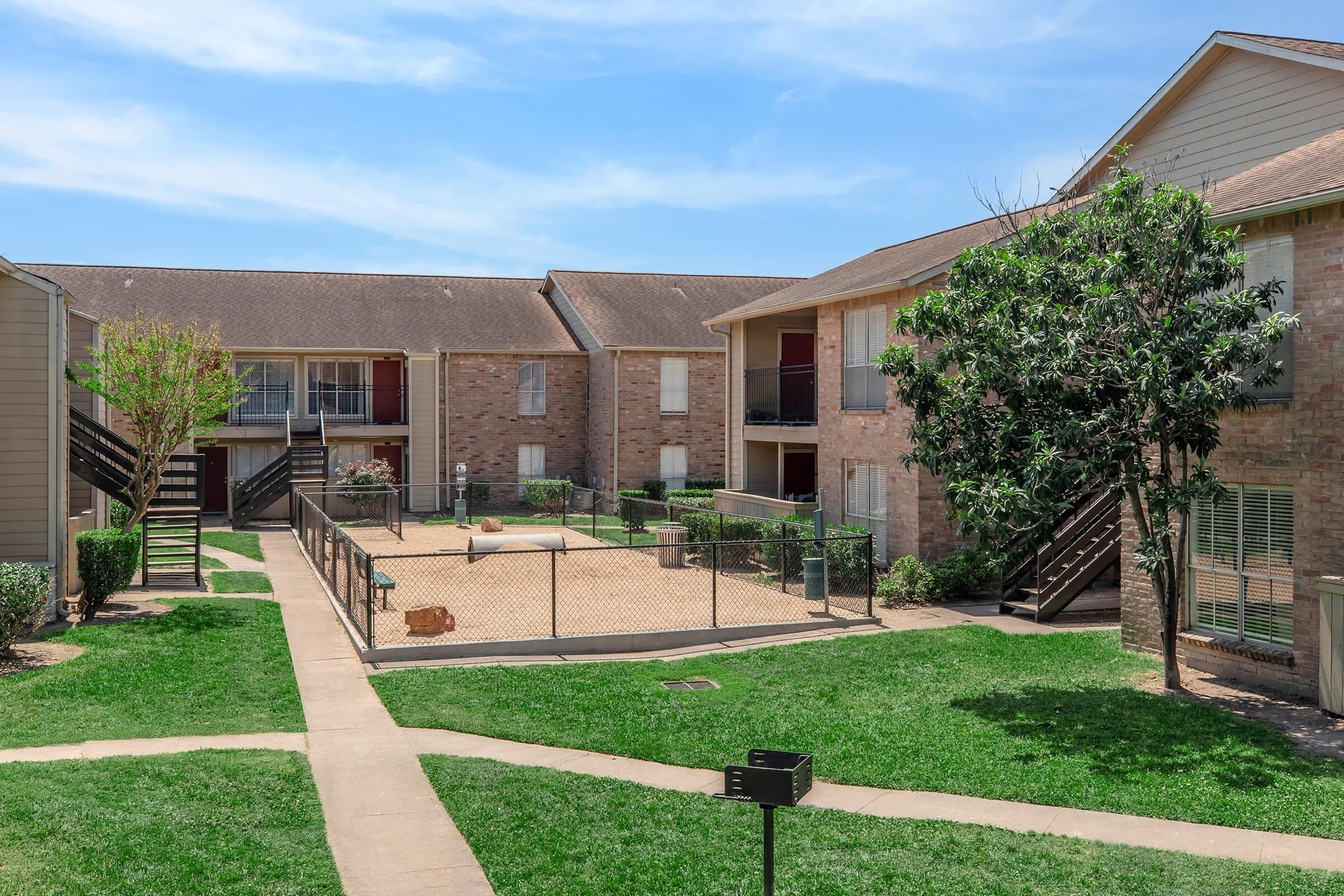 A well-maintained apartment complex featuring two-story buildings surrounding a central courtyard with green grass. A fenced dog park is visible in the courtyard, along with trees and walkways. The blue sky above adds to the pleasant ambiance.
