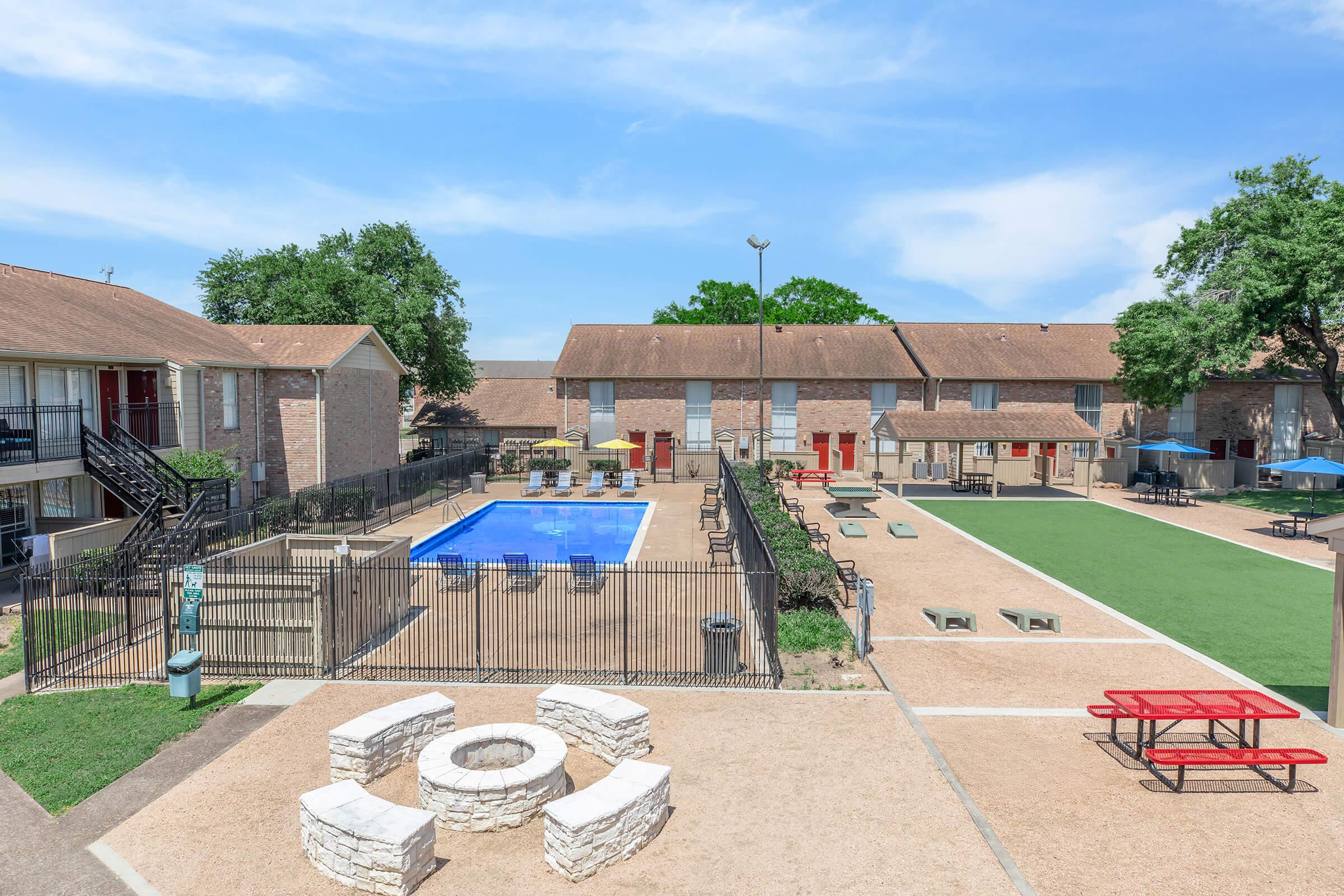 A view of an apartment complex featuring a blue swimming pool surrounded by lounge chairs and picnic tables. The area includes a grassy space, barbecue grills, and shaded seating under umbrellas. The buildings are two stories, with staircases and railings visible. The sky is clear and blue.