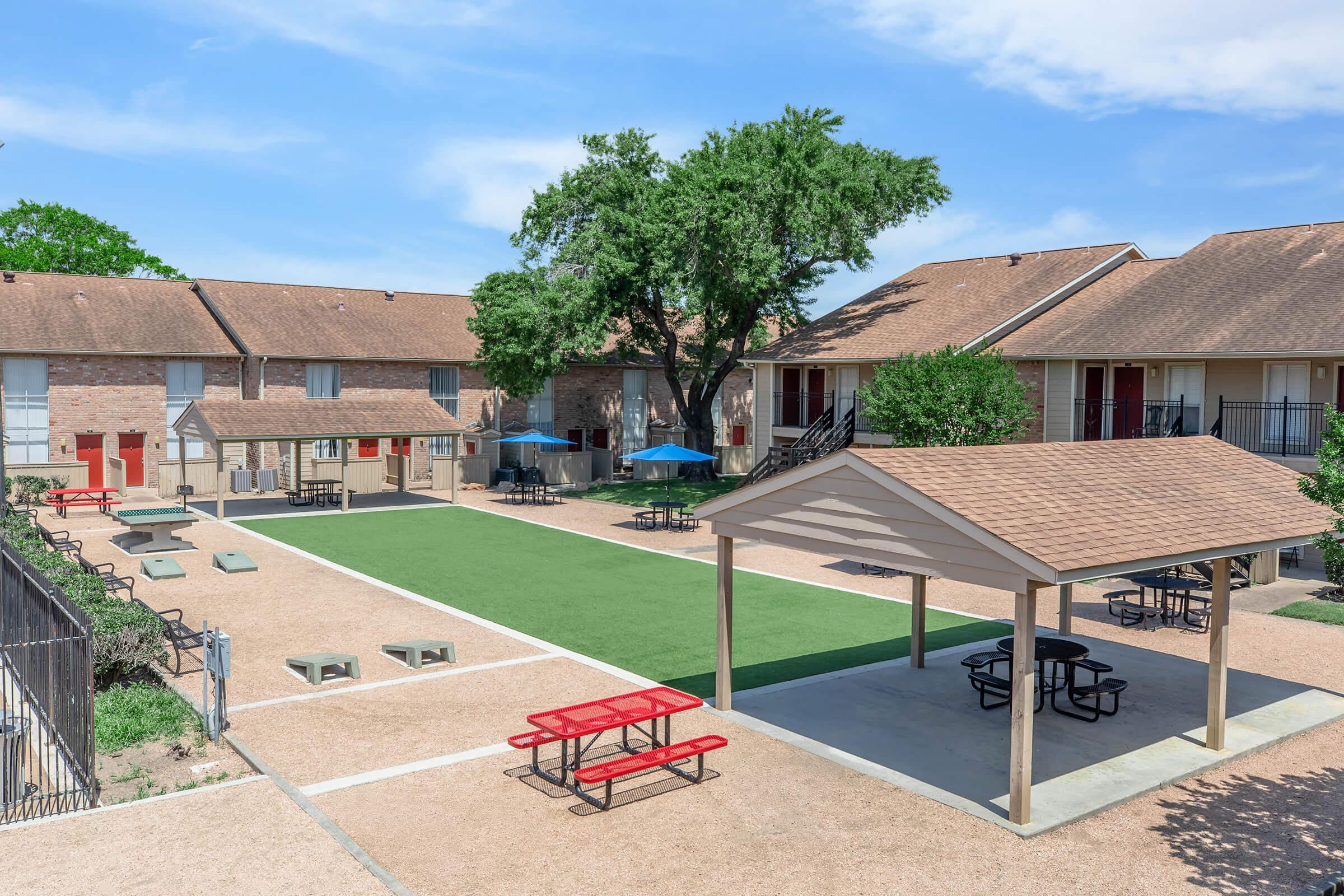 A courtyard featuring green turf, surrounded by residential buildings. Picnic tables are arranged beneath a shaded pavilion, with several blue umbrellas providing shade. The area is well-maintained and includes walkways and trees, creating a welcoming outdoor space for residents.