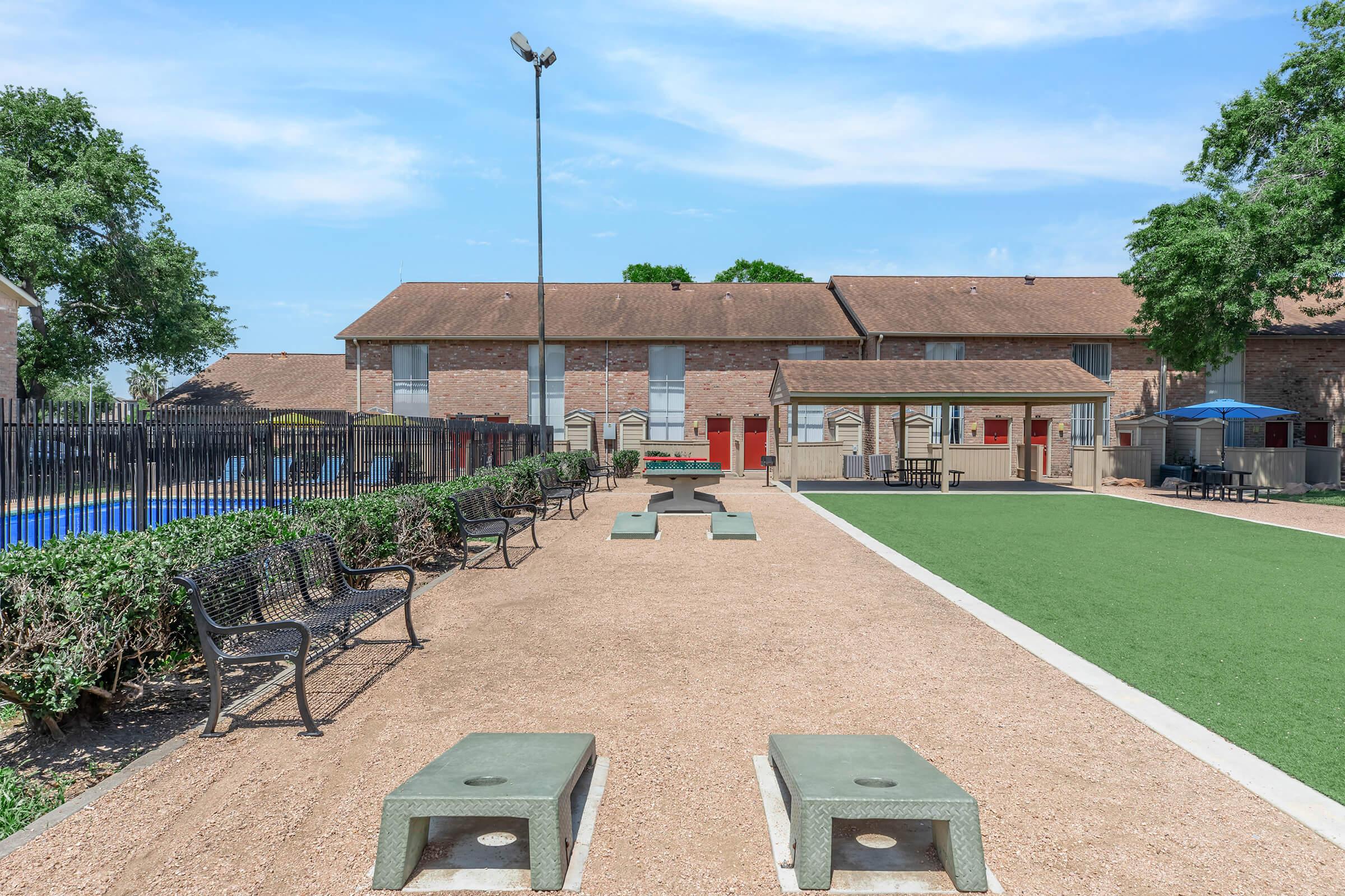 A spacious outdoor recreational area featuring benches and seating, surrounded by neatly trimmed hedges. In the background, there are buildings with red doors. The area includes a grassy lawn and a pool area, all under a clear blue sky.
