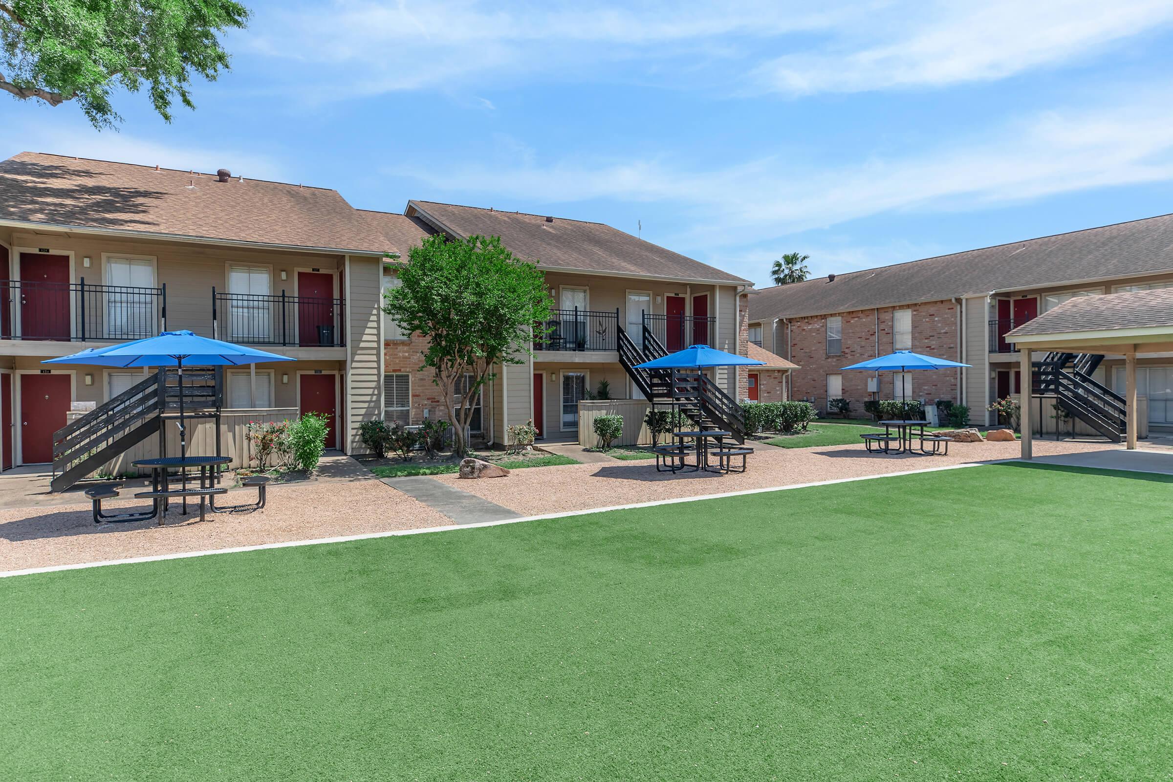 View of an apartment complex featuring two-story buildings with balconies, surrounded by green artificial turf. There are several blue umbrellas over picnic tables and walking paths, with trees providing shade. The sky is clear and blue, contributing to a pleasant outdoor atmosphere.