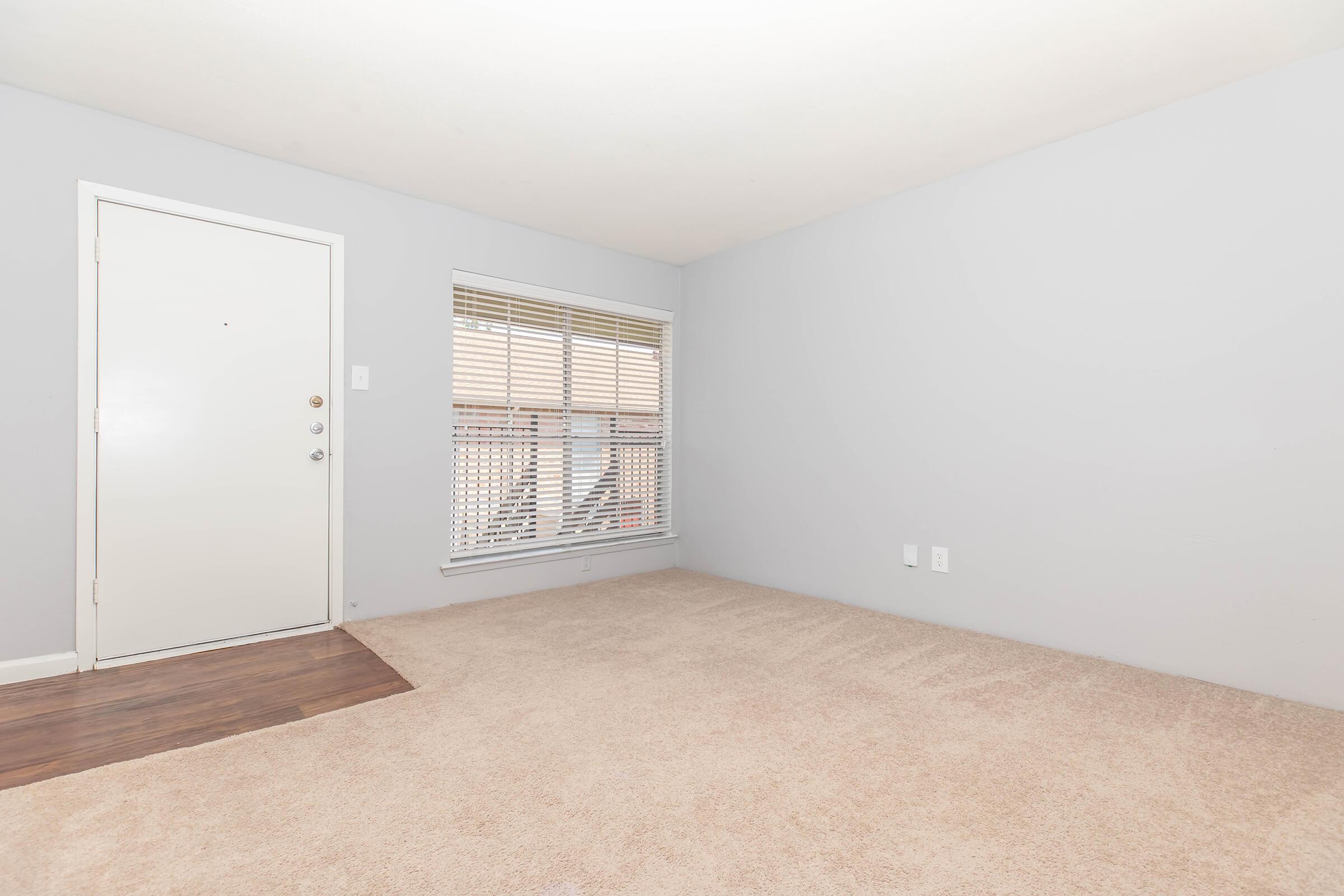 Empty room with light gray walls and a beige carpet. A closed white door is on the left, and a window with horizontal blinds is positioned on the right. The overall atmosphere is bright and spacious, with minimal furniture or decor present.