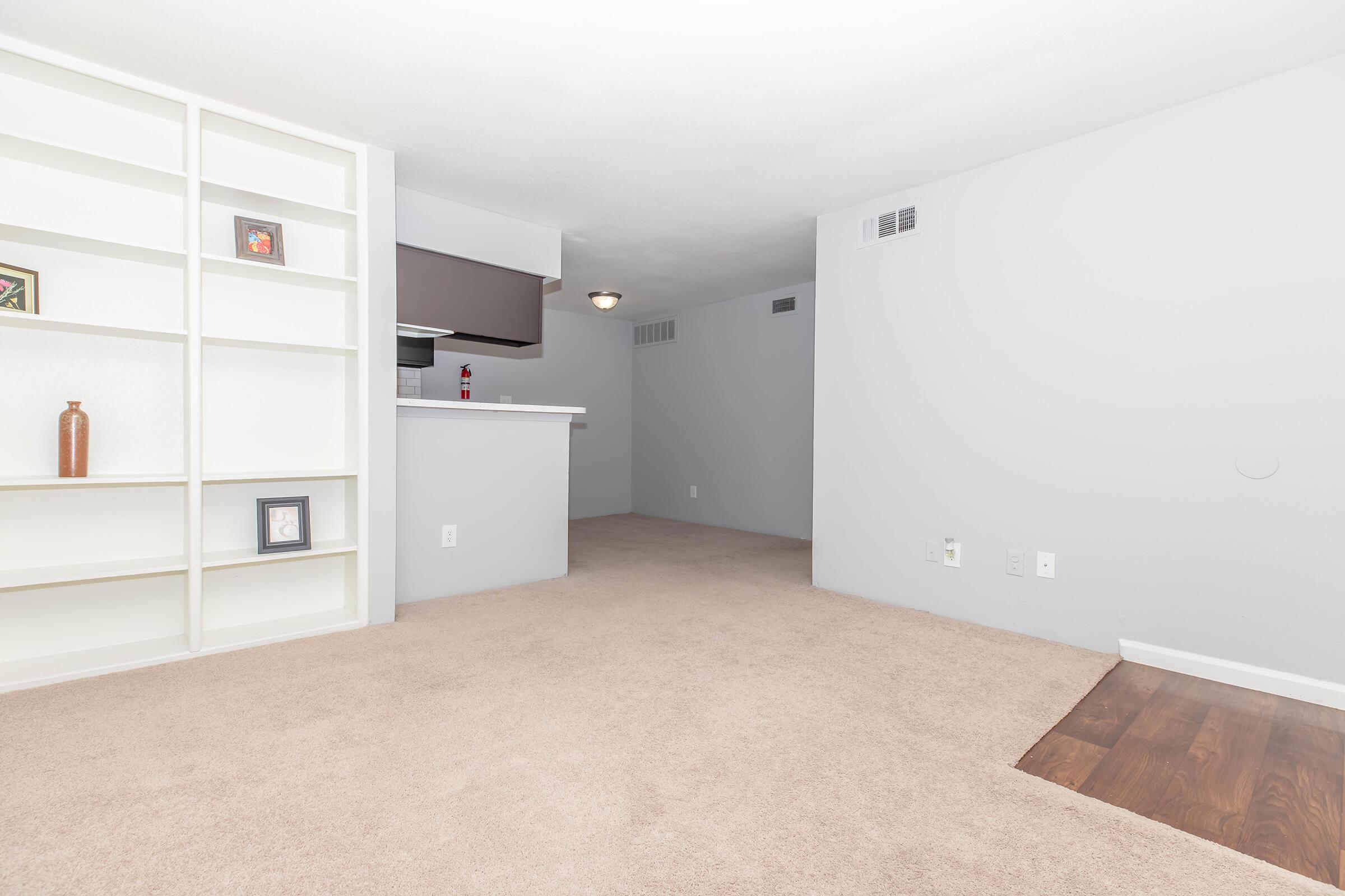 A spacious and neutral-toned living area featuring beige carpet, a built-in white bookshelf with decorative items, and an open view to a modest kitchen area. Soft lighting enhances the inviting atmosphere, while light grey walls contribute to a modern and minimalist design.