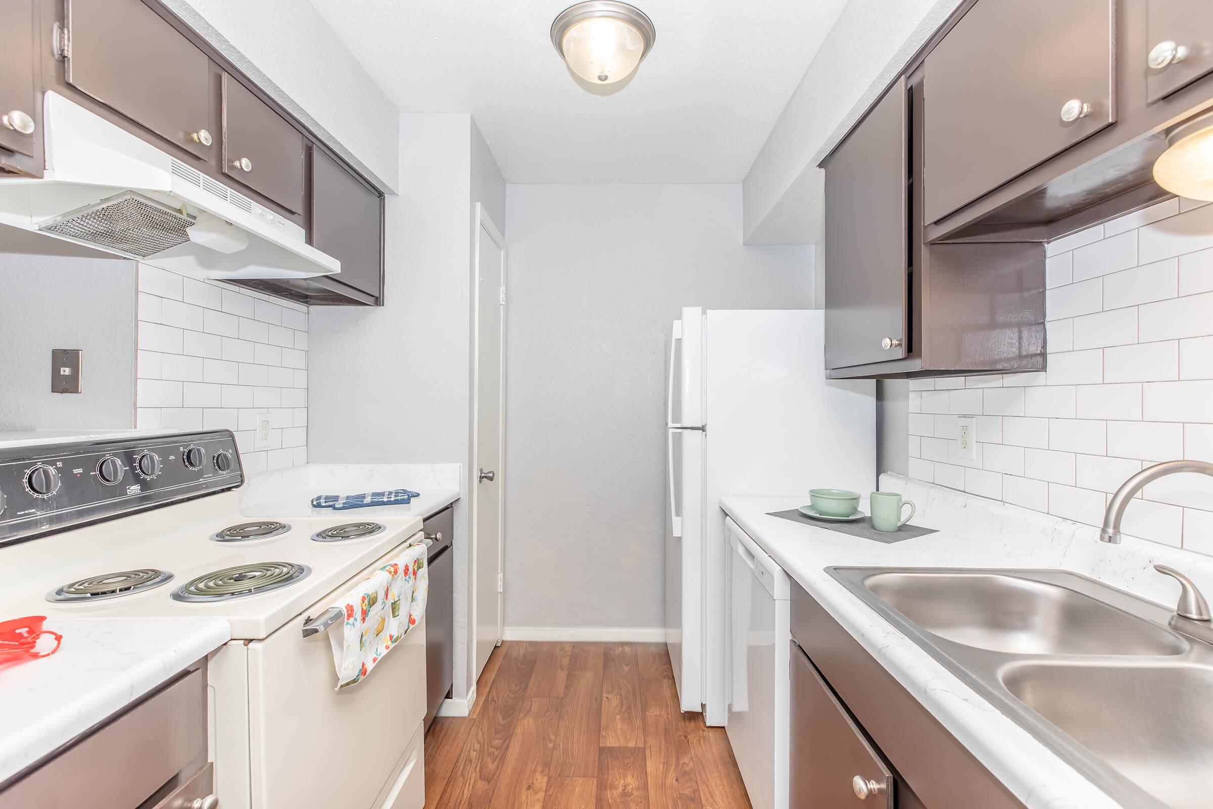 A modern kitchen featuring dark wood cabinets, a white tiled backsplash, an electric stove, and a double sink. There are stainless steel appliances, including a fridge, and wooden flooring. A small counter space is visible, with a towel and two green cups placed on it.