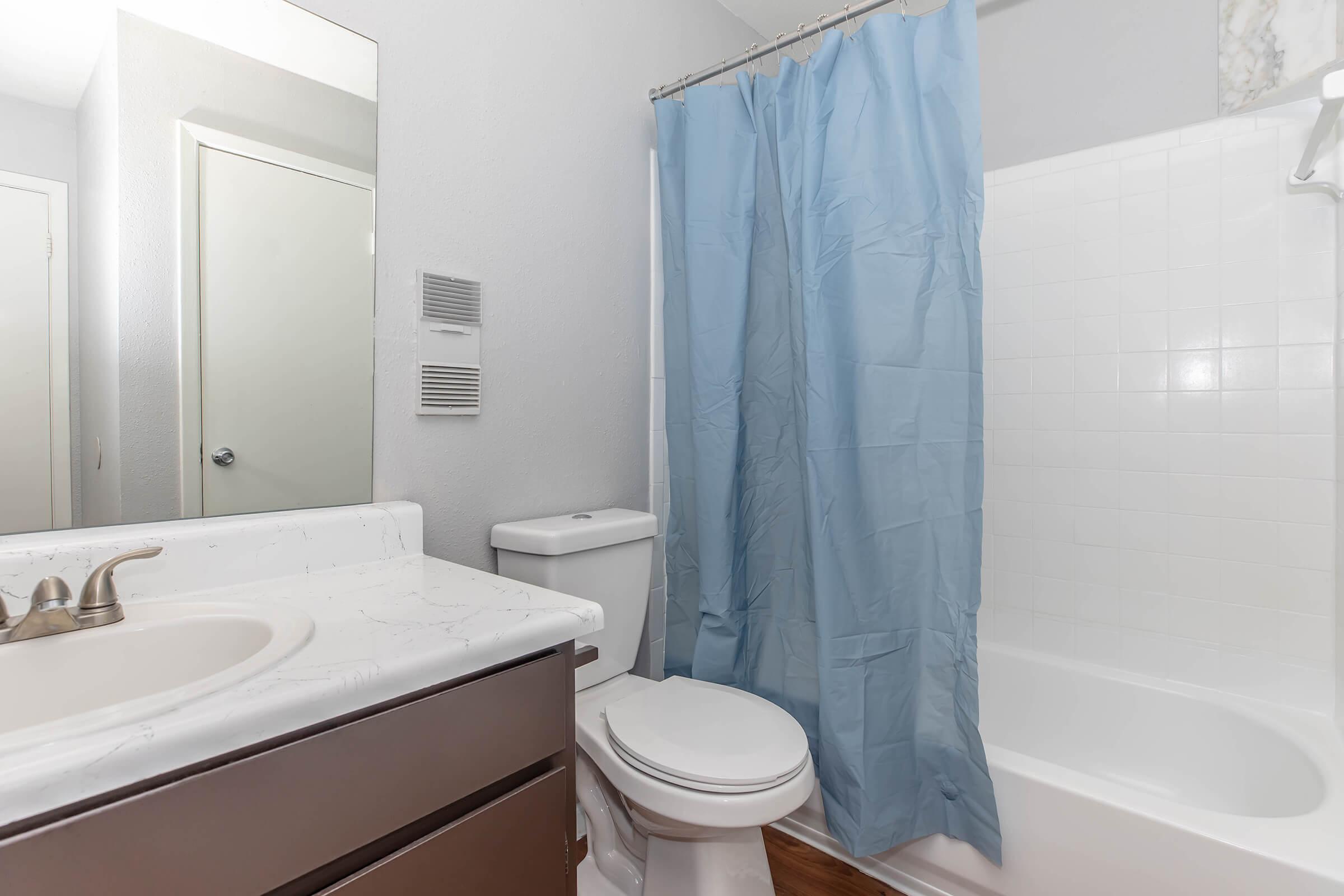 A clean bathroom featuring a white bathtub and shower combination, a light brown vanity with a sink, a toilet, and a blue shower curtain. A mirror is mounted above the vanity, and the walls are painted light gray. The floor has wood-like tiles, creating a modern and fresh appearance.