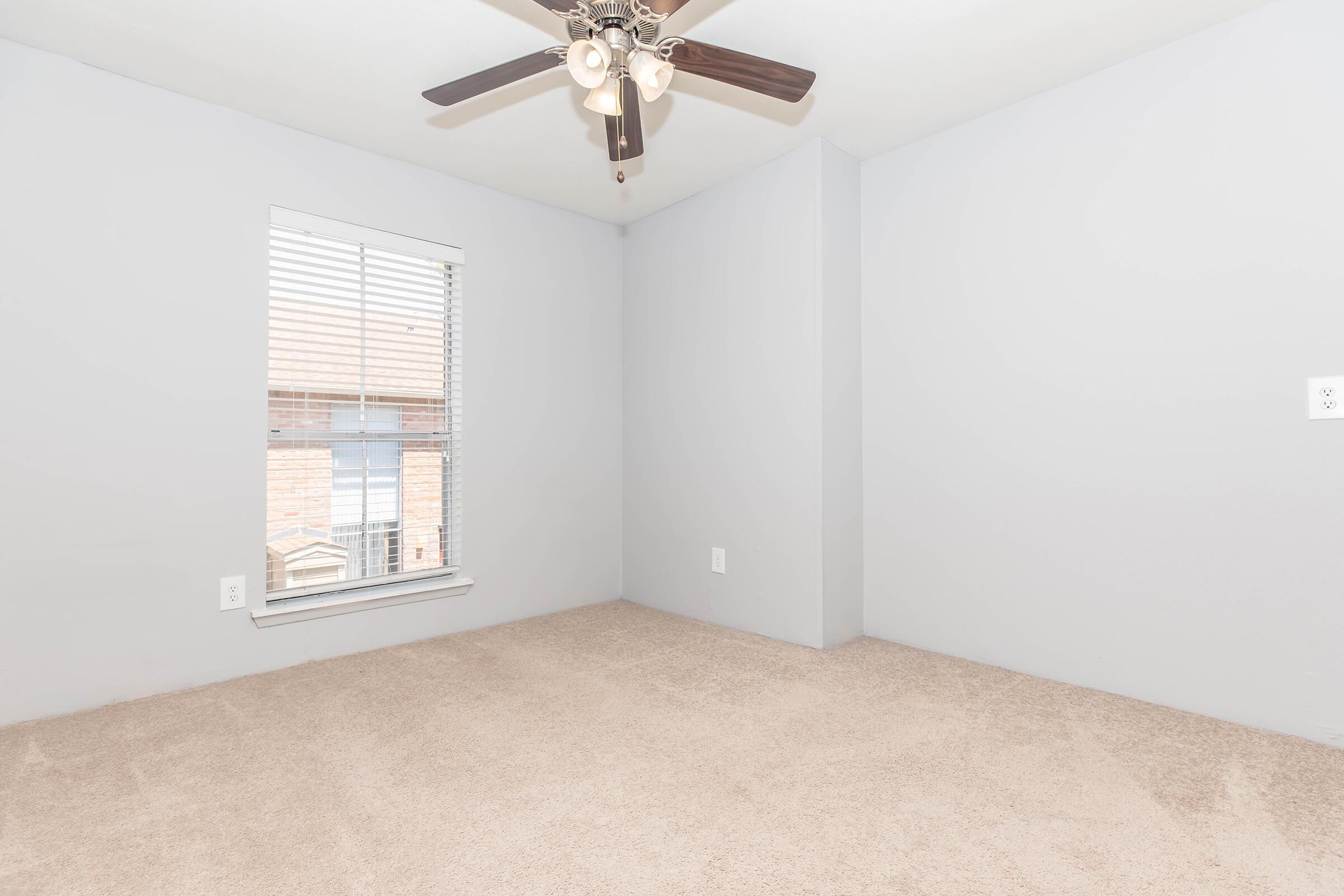 Empty room with light gray walls and a ceiling fan. A window with blinds allows natural light to enter, illuminating the soft beige carpeted floor. The room appears spacious and ready for furnishing.