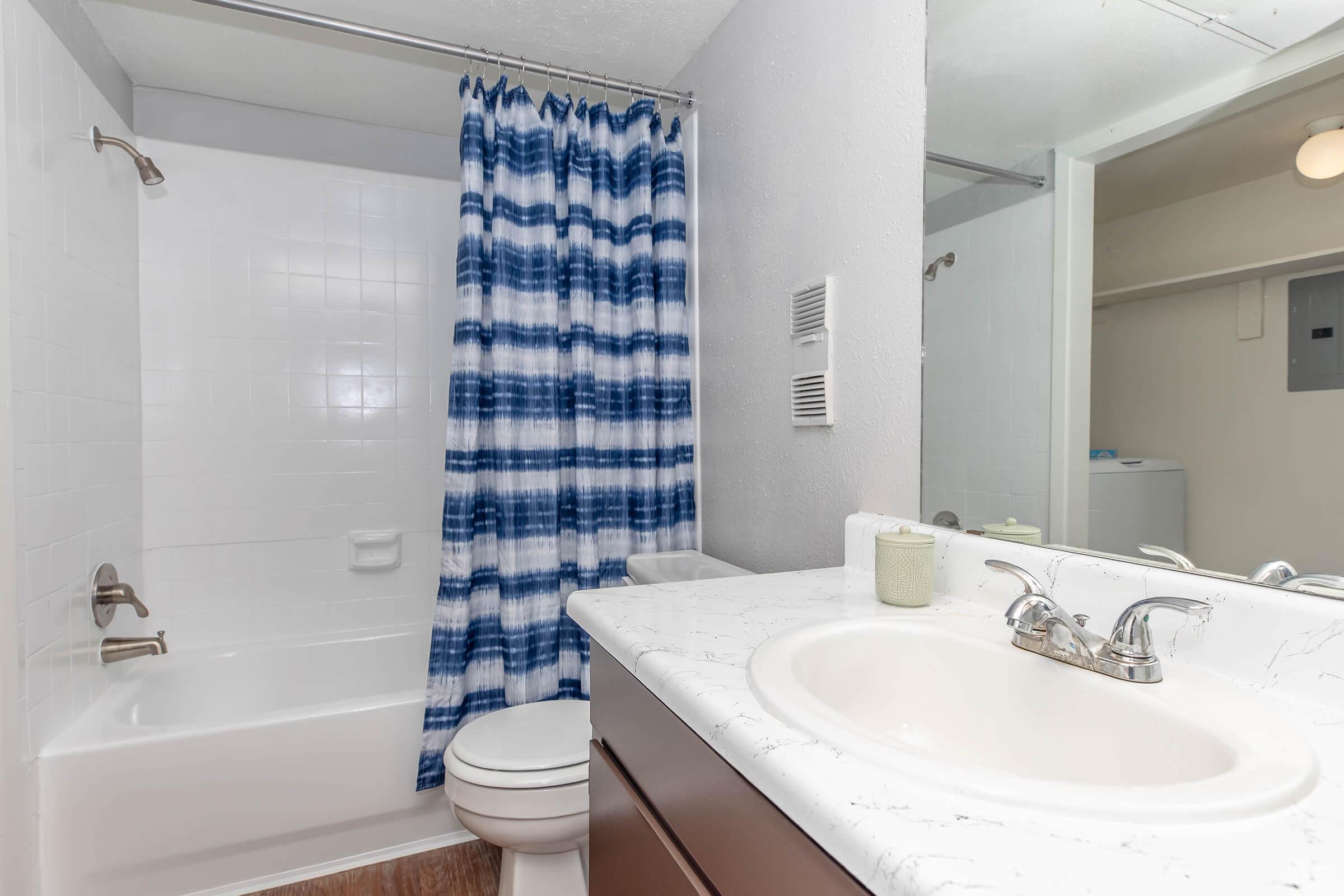 A modern bathroom featuring a white bathtub with a blue and white striped shower curtain, a white sink with a marble countertop, and a wooden cabinet. There are light-colored walls, and the space appears clean and tidy, with a glimpse of laundry facilities in the background.