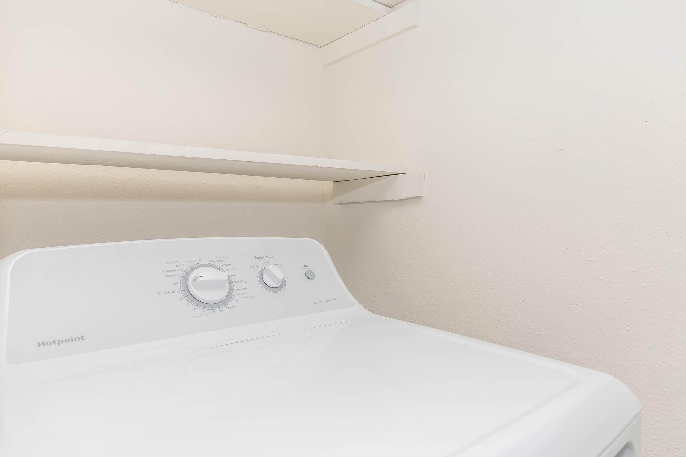A white Hotpoint washing machine is situated in a minimalistic laundry space with beige walls. There is a shelf above the machine, and the overall environment appears tidy and simple.
