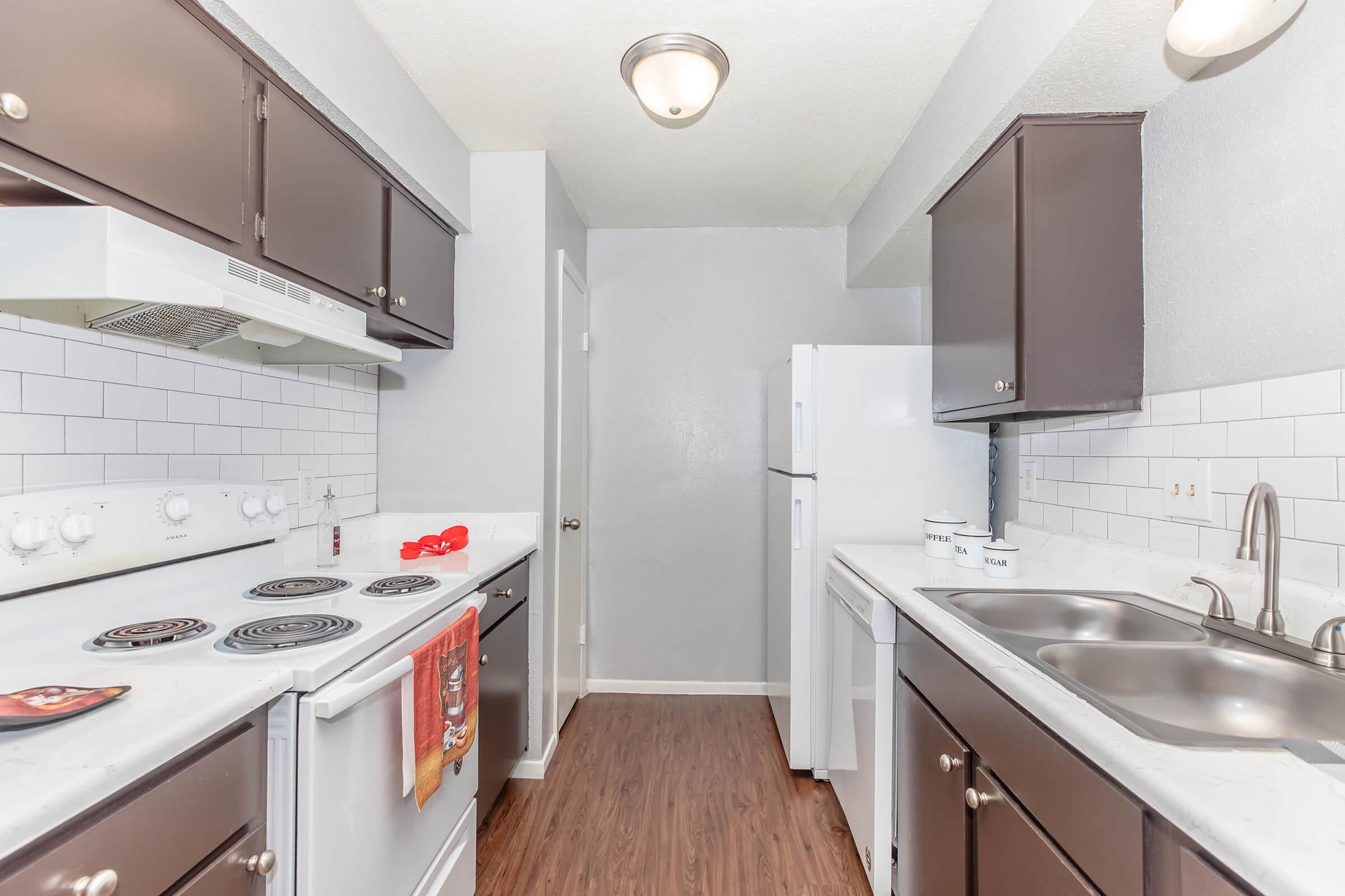 A modern kitchen featuring dark cabinets, a white stove and oven, a double sink, and stainless steel appliances. The walls are painted in a light gray, and the flooring resembles dark wood. There are white subway tiles as a backsplash, and a small red towel hangs from the oven.