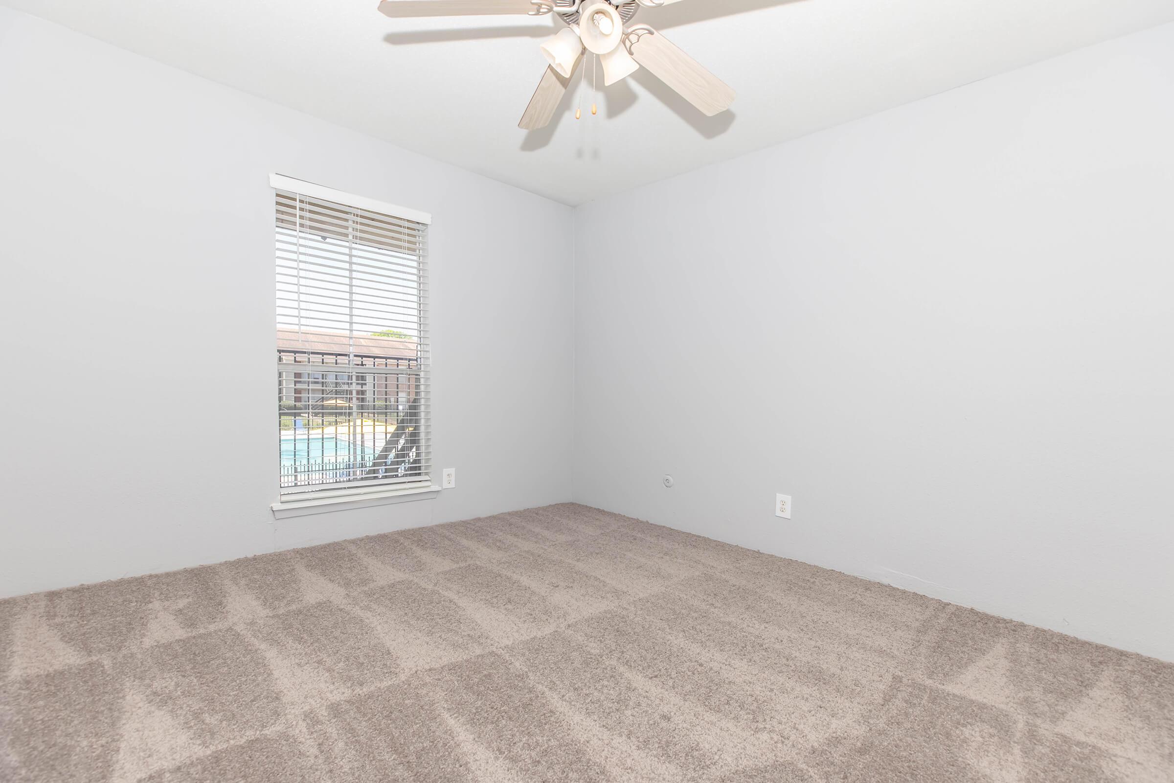A sparsely furnished room with light gray walls and carpet. A ceiling fan is visible, and there is a window with blinds that overlooks a pool area. The room is empty, creating an open and airy feel.