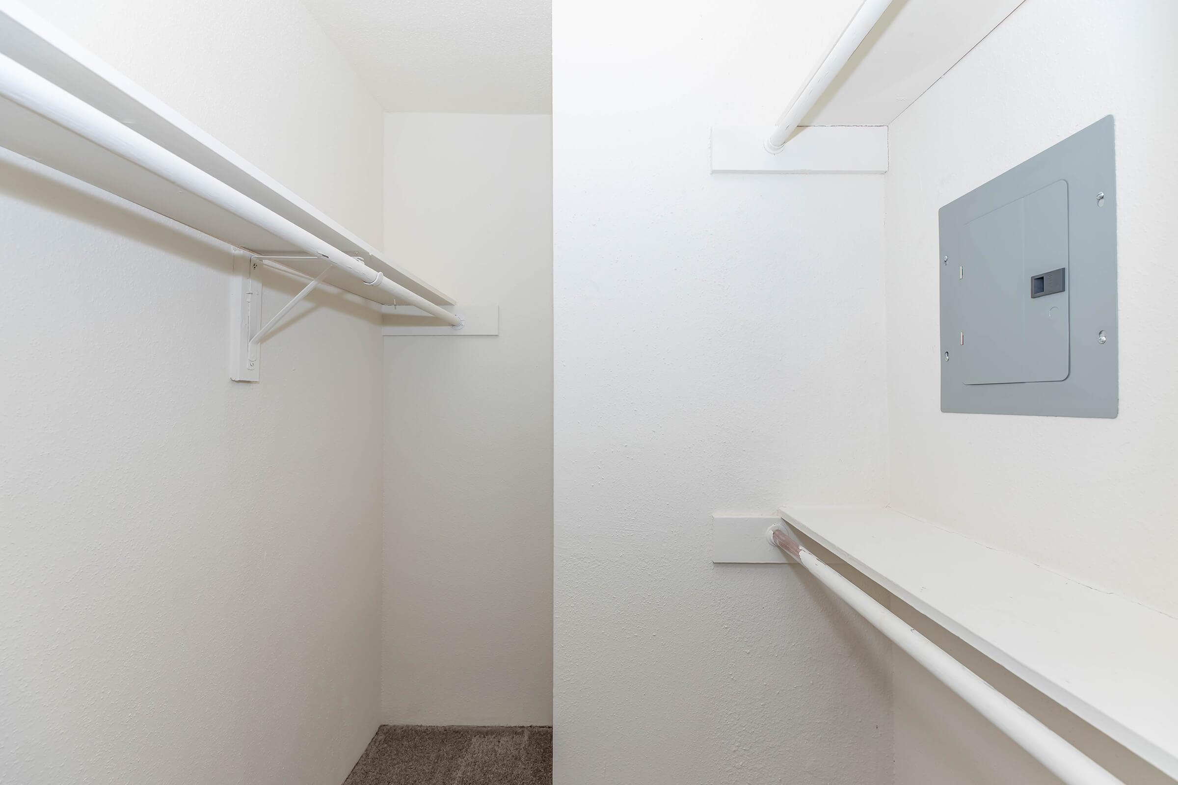 An empty closet with white walls and a gray electrical panel. There are two clothes hanging rods made of white piping on opposite sides of the closet. The floor is carpeted in a neutral color. The overall appearance is clean and minimalistic.