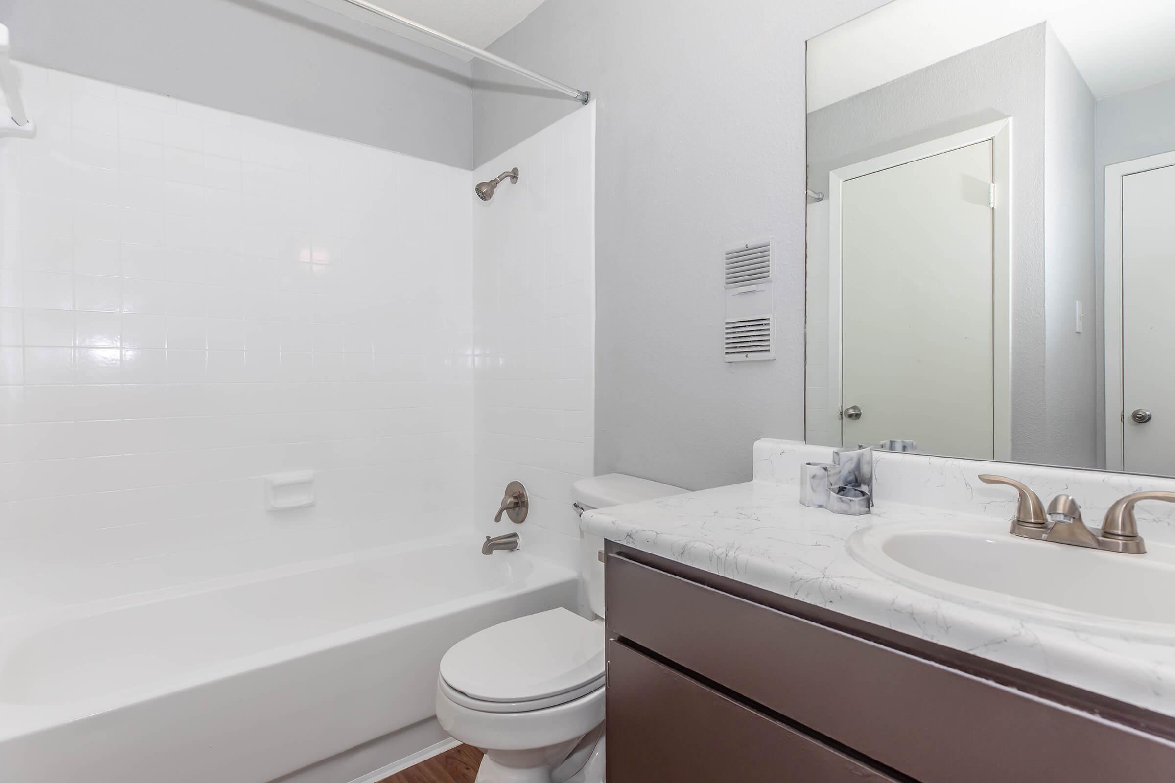 A clean, modern bathroom featuring a bathtub with a shower, a white toilet, and a marble countertop sink. The walls are light gray, and there is a large mirror above the sink. A door leads to another room, and there are air vents on the wall. Natural light may enter from outside the frame.