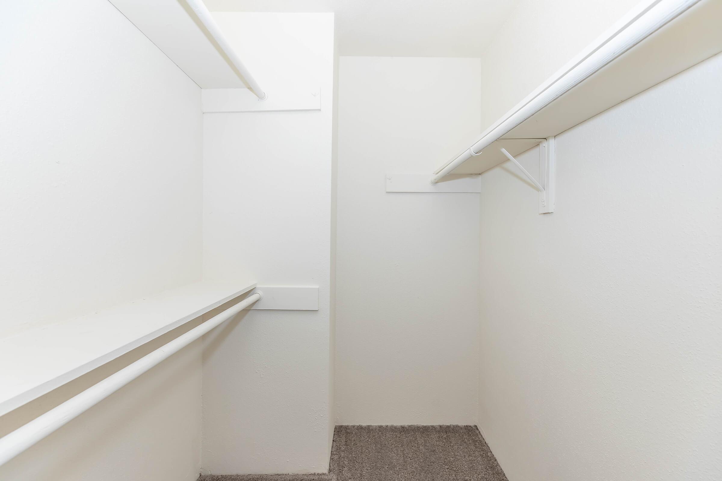 An empty closet with white walls and two horizontal shelving units. The floor is covered with gray carpet. The closet has a clean and minimalistic design, with ample space for storage.