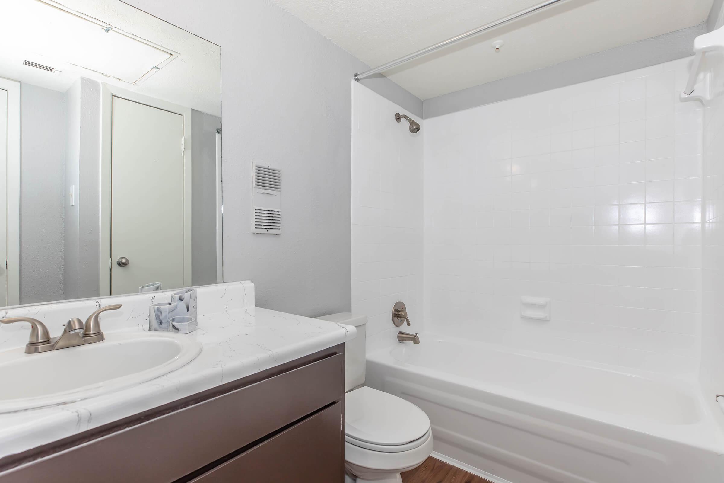 A clean, modern bathroom featuring a white bathtub with a shower, a large mirror above a marble countertop, and a toilet. The walls are painted light gray, and the floor has wooden laminate. A bathroom door is partially visible in the background.