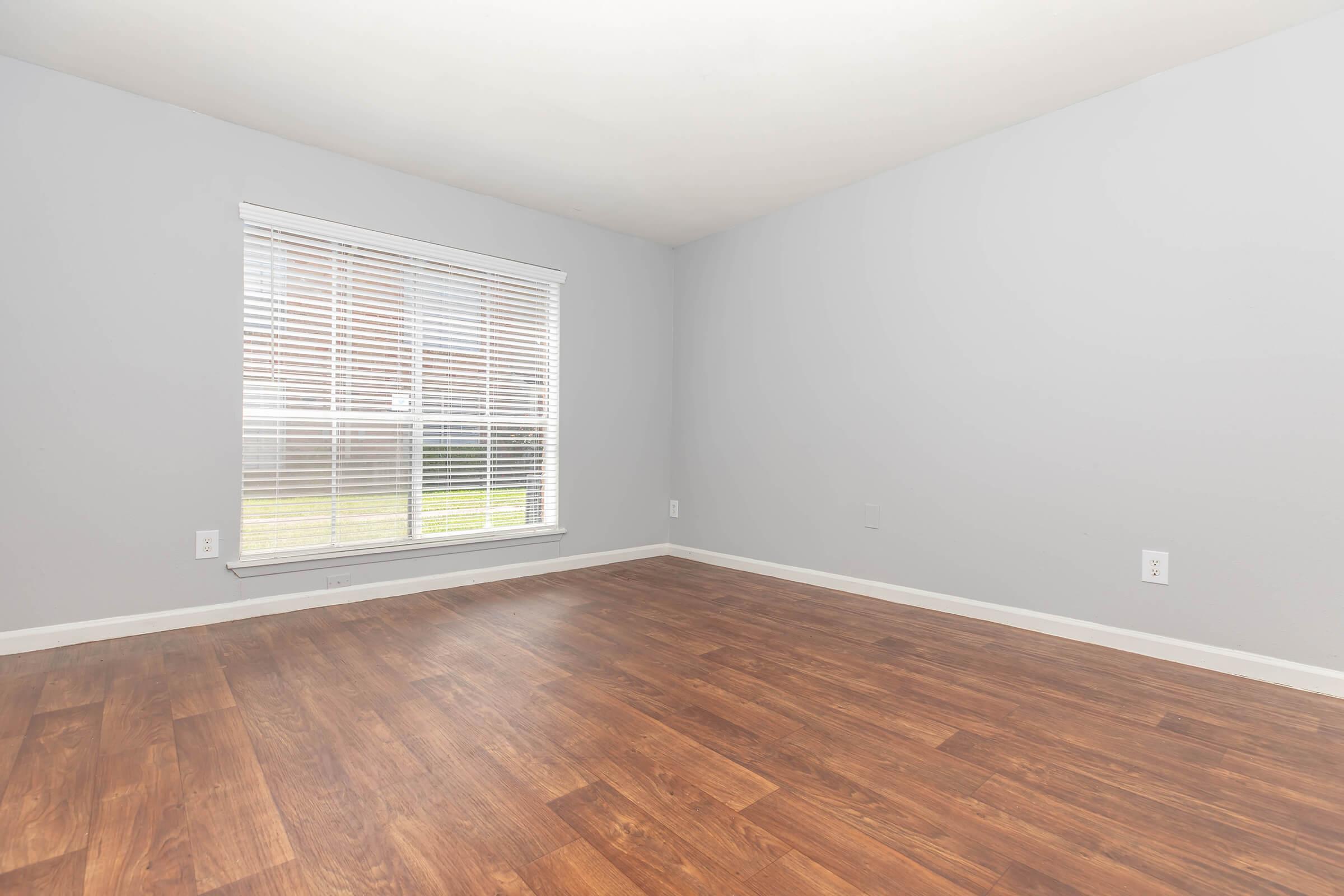 Empty room with light gray walls and a large window covered by white blinds. The floor is made of brown laminate wood, and there are no furnishings or decorations, creating a clean and spacious appearance. Natural light is coming in from the window, illuminating the space.