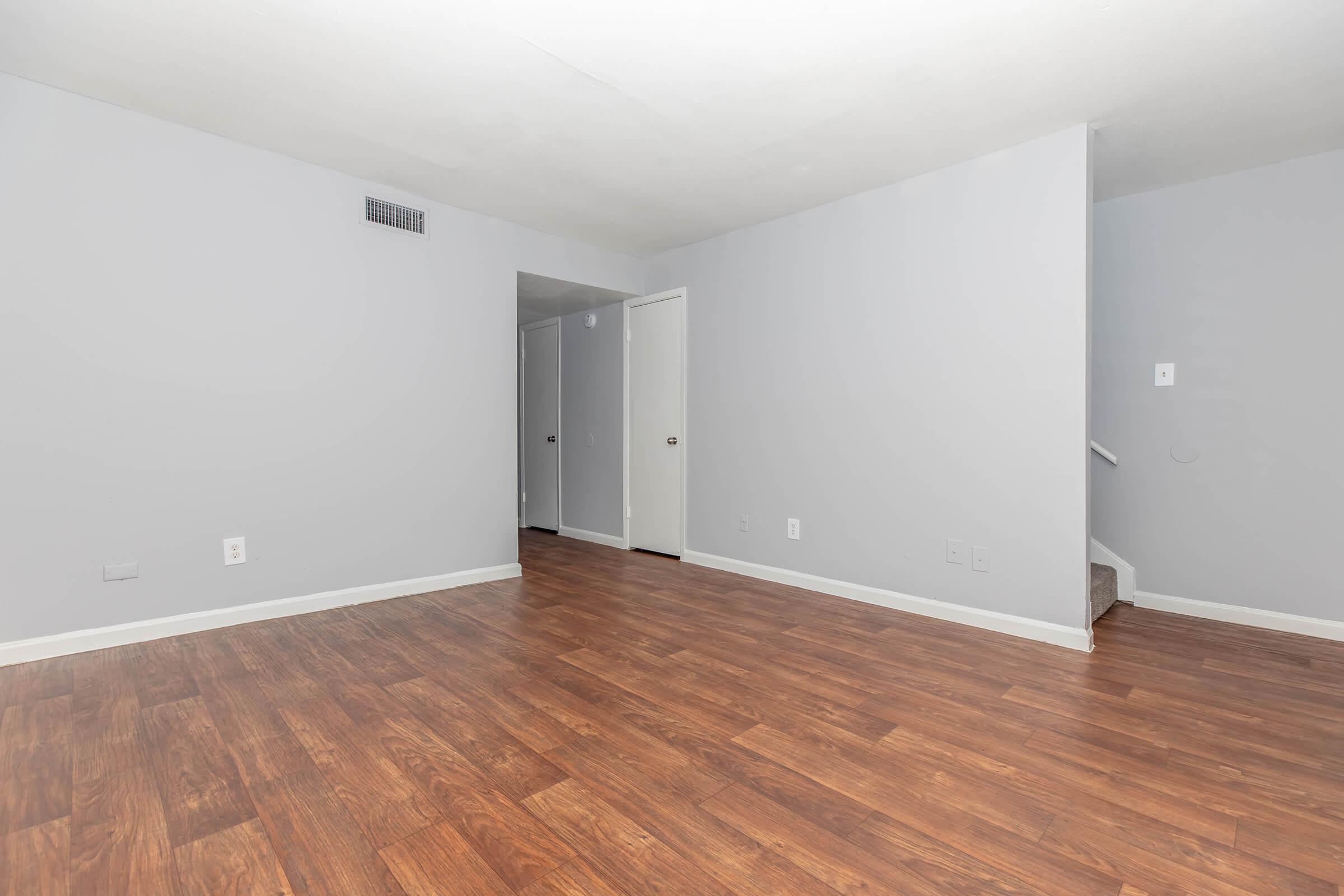 A spacious, empty room with light gray walls and a dark wood floor. A doorway on the right leads to a staircase, and there are two closed doors on the left side of the wall. The area appears well-lit and ready for furnishing.