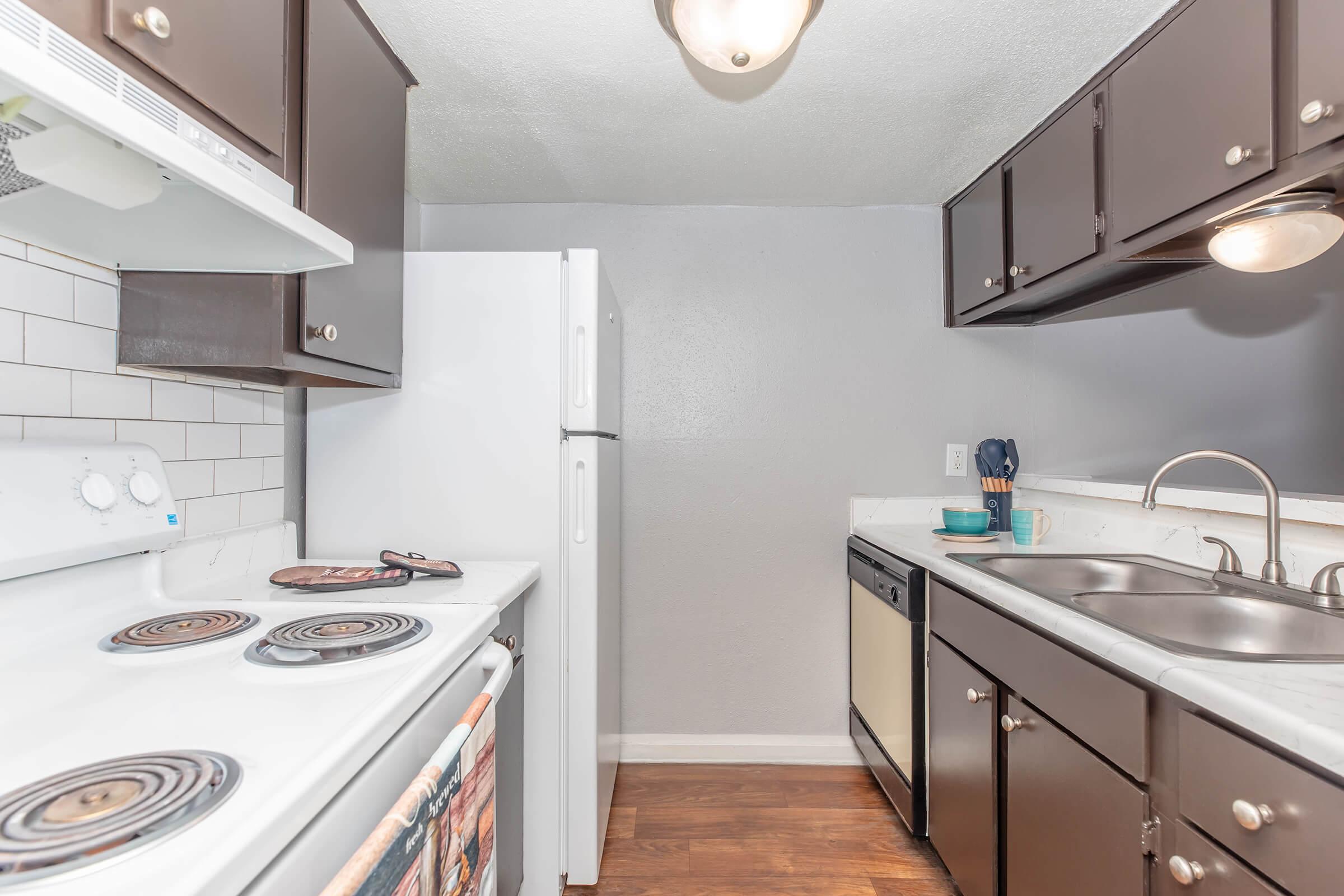 A modern kitchen featuring dark wood cabinets, a white stove and refrigerator, and a double sink. The countertops are light-colored, and there's a dishwasher. The walls are painted in a neutral tone, creating a clean, functional space.