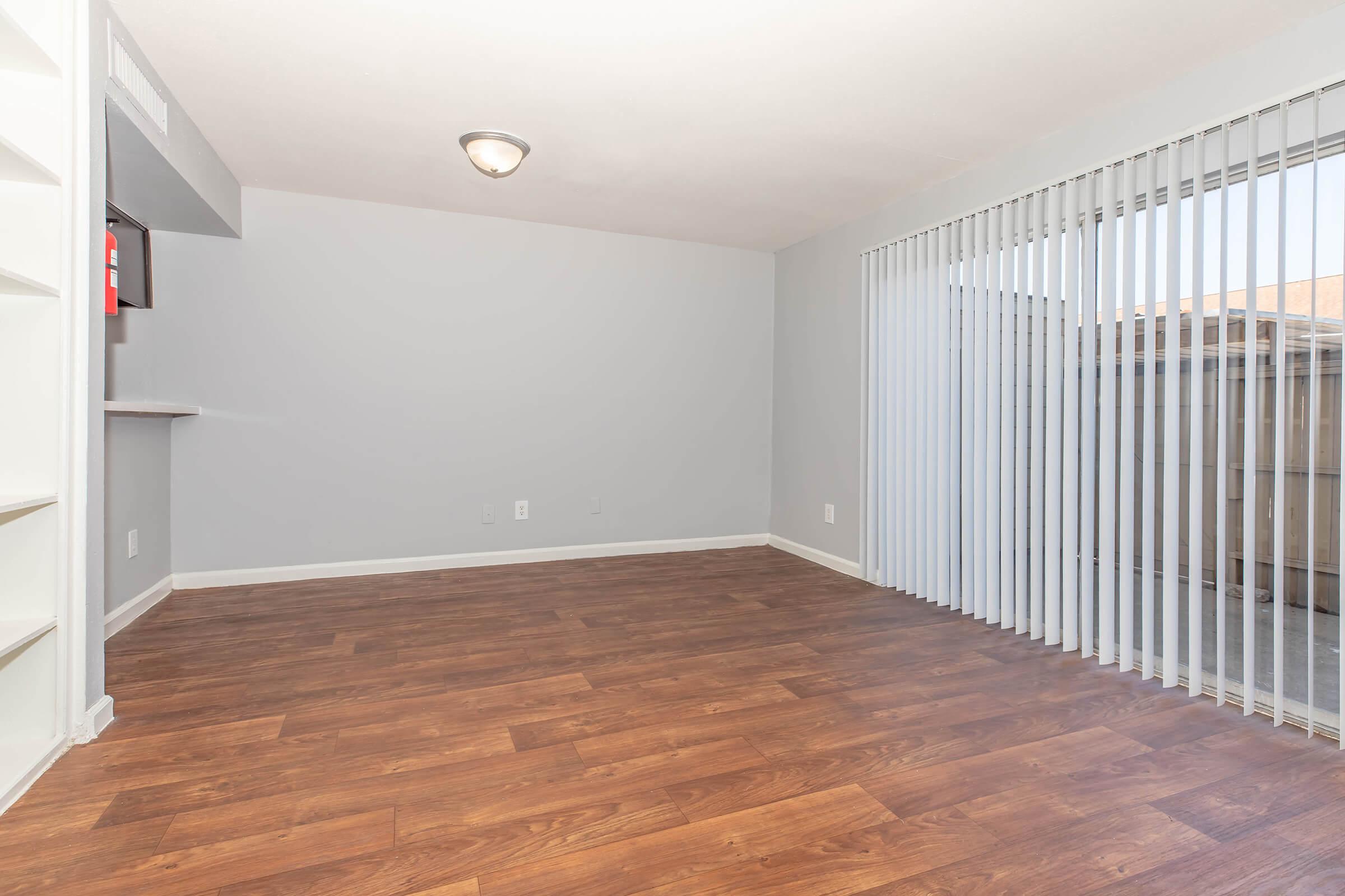 Empty indoor living space featuring light gray walls, a ceiling light fixture, and wooden laminate flooring. A sliding glass door with vertical blinds leads outside, while an open kitchen area is visible in the background. The room is well-lit and spacious, suitable for various furniture arrangements.