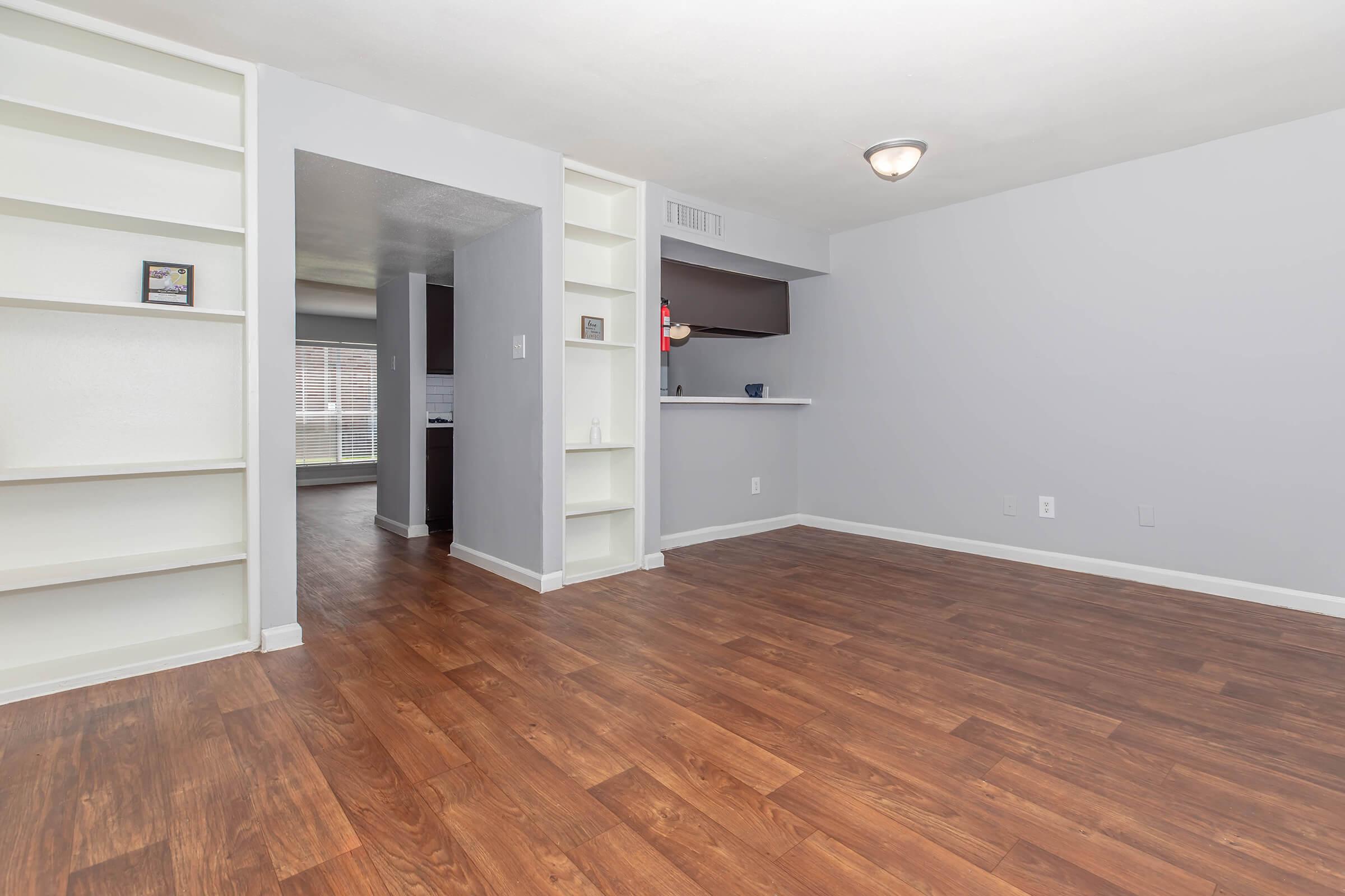 Spacious interior of a modern apartment with laminate flooring, light gray walls, and built-in shelves. View into an adjacent room through an opening, featuring a kitchen area in the background. Natural light filters in through a large window, creating a welcoming atmosphere.