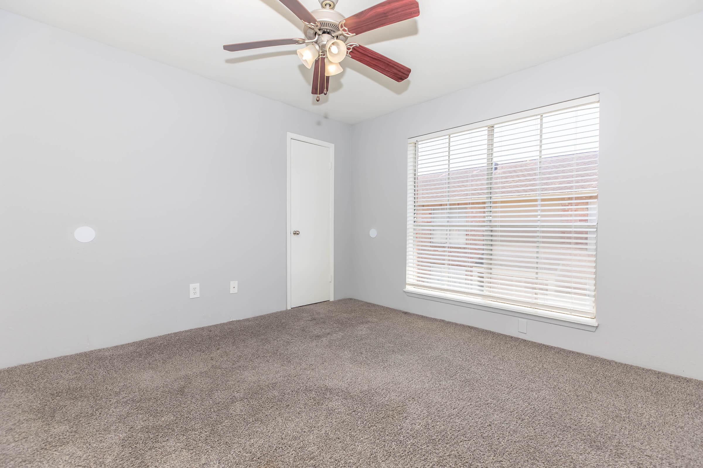 An empty room with light gray walls and a ceiling fan with brown blades. There is a closed white door on one side and a large window with horizontal white blinds allowing natural light to enter. The floor is covered with light brown carpet.