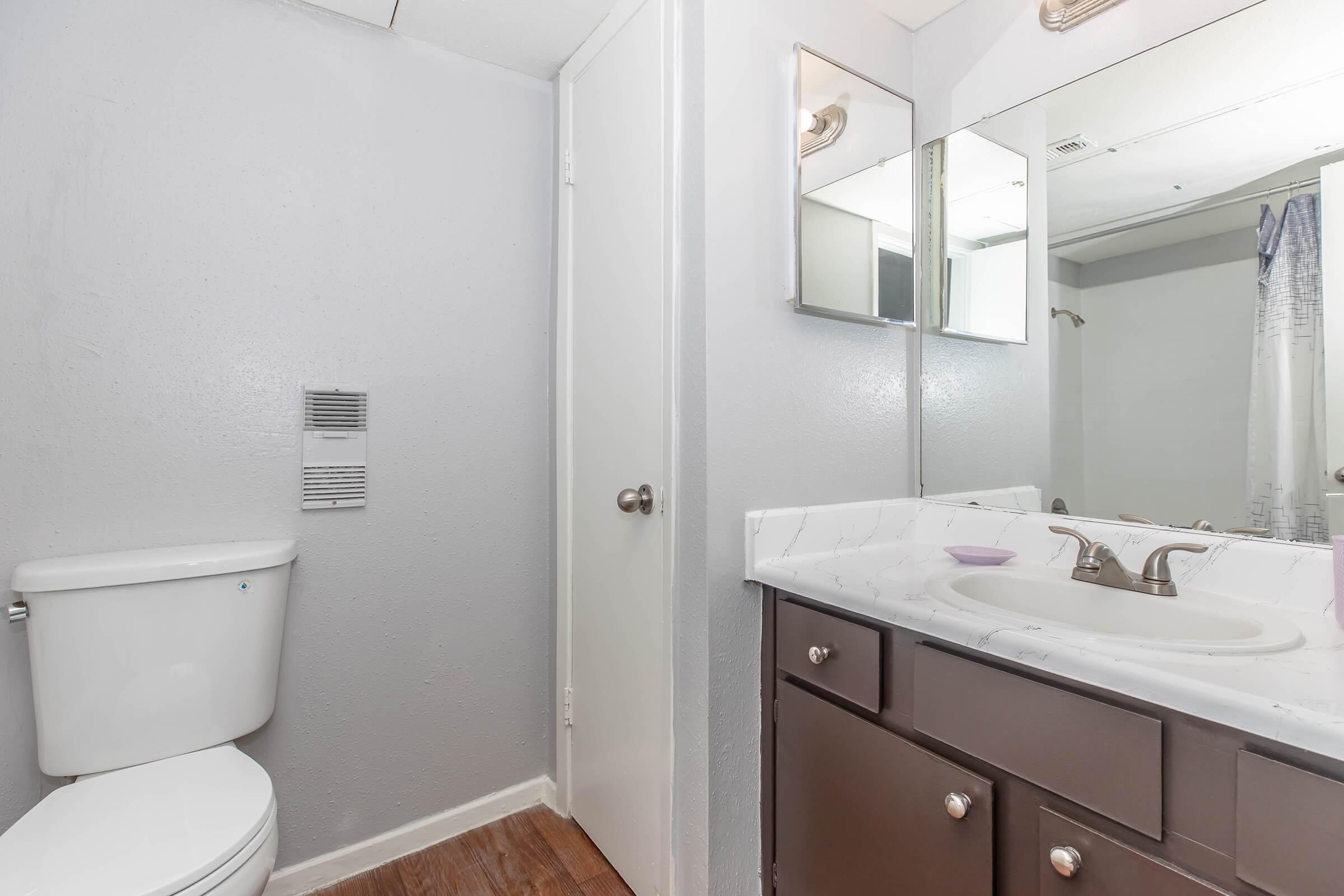 A clean, modern bathroom featuring a white toilet, a sink with a countertop, and two mirrors above the sink. The walls are painted in a light gray, and there's a door leading to another area. The flooring is brown, and a shower curtain is visible in the background.