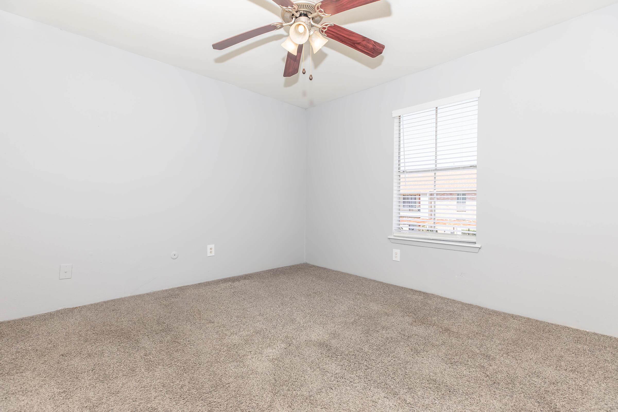 Empty room with light gray walls and a ceiling fan. The floor is carpeted in a beige tone. A window is visible on the right side, allowing natural light to enter. The room has no furniture or decor, creating a spacious and minimalistic appearance.