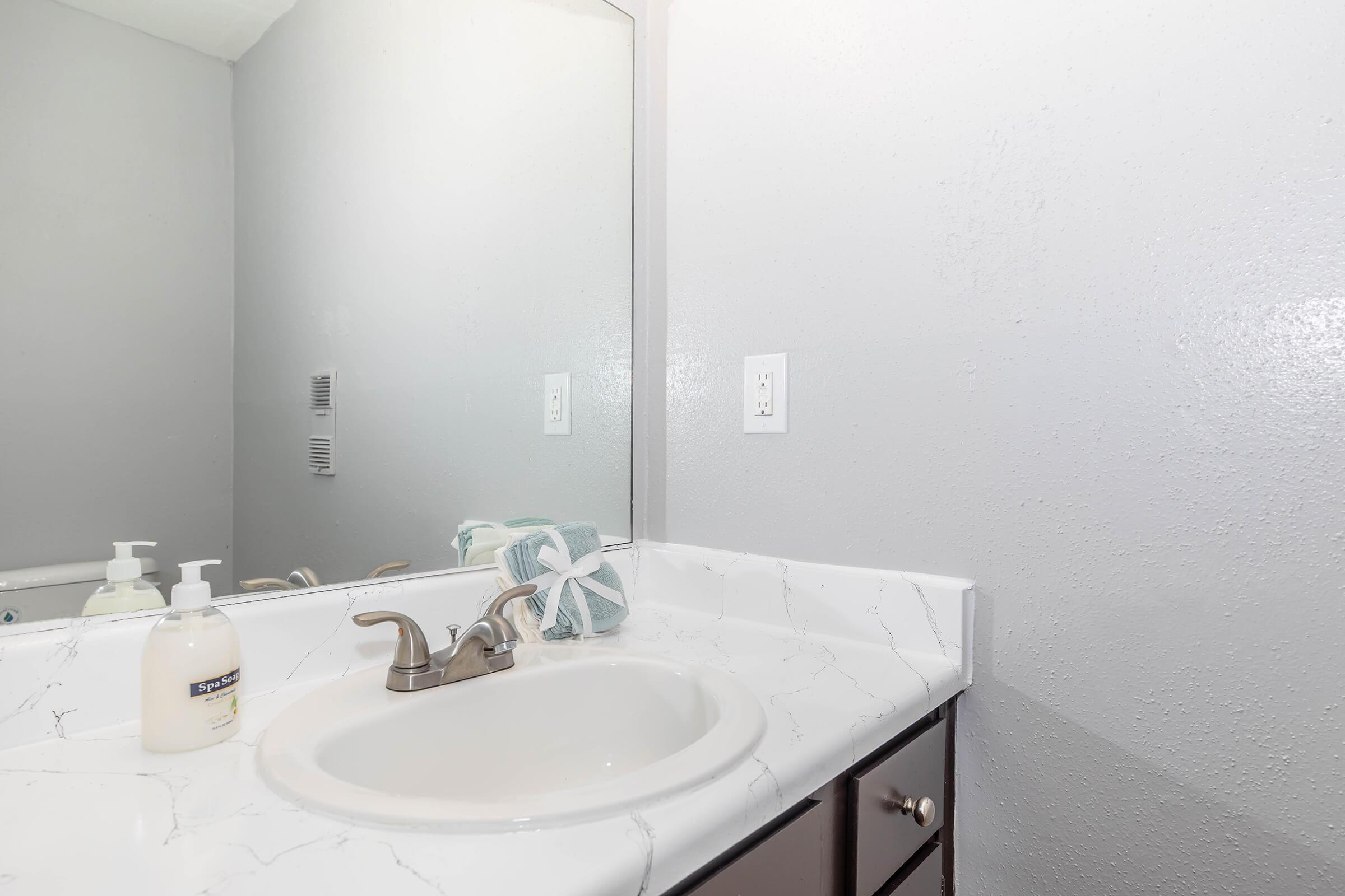 A clean bathroom sink area featuring a white countertop, a silver faucet, and a mirror. Two bottles of soap are placed next to a hand towel on the counter. The wall is painted in a light gray color, creating a simple and modern aesthetic.