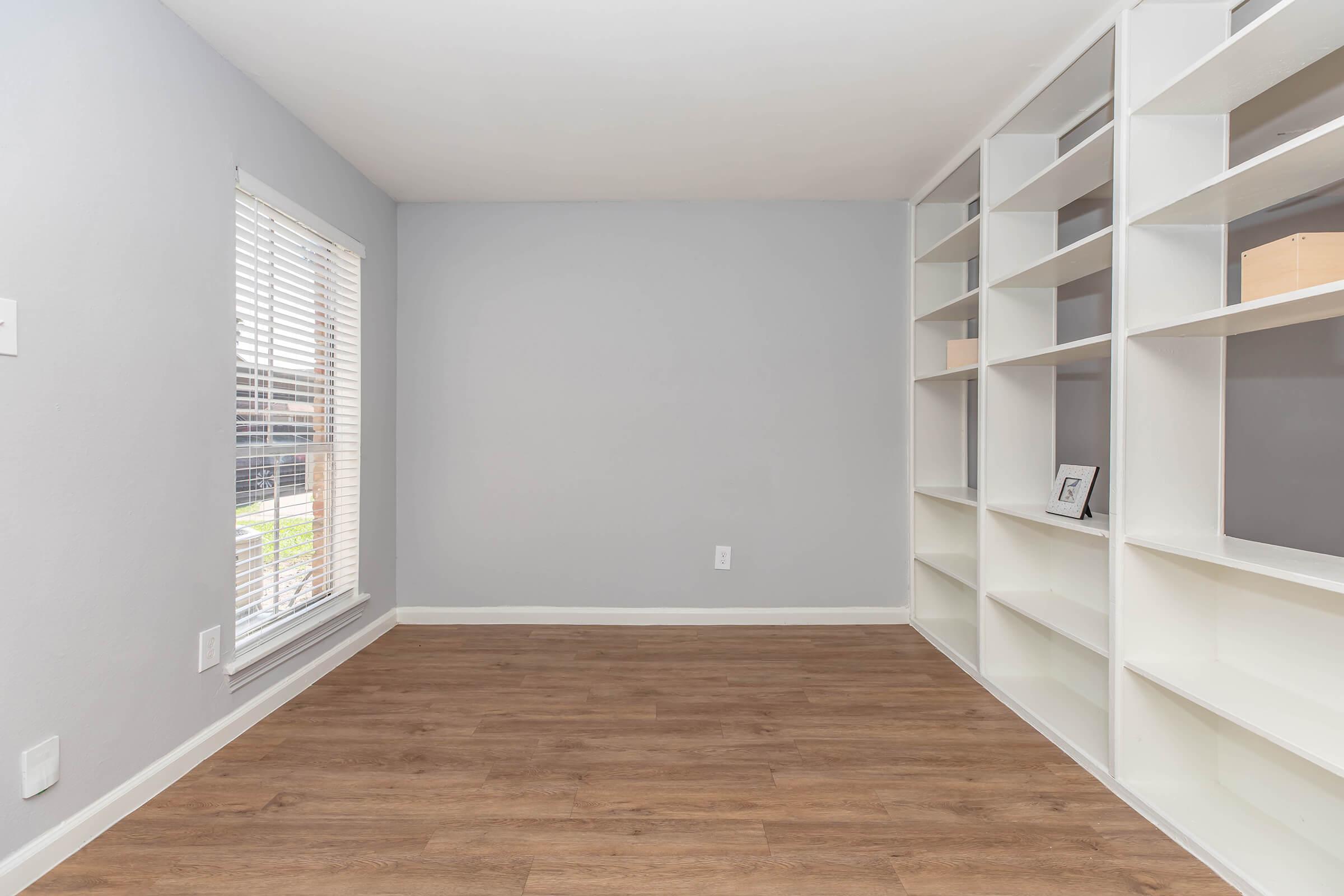 A minimalistic room with light gray walls and hardwood flooring. There is a large window with white blinds allowing natural light to enter. On one wall, there is a white shelving unit with open shelves. The space is staged to appear open and airy, with no furniture present.