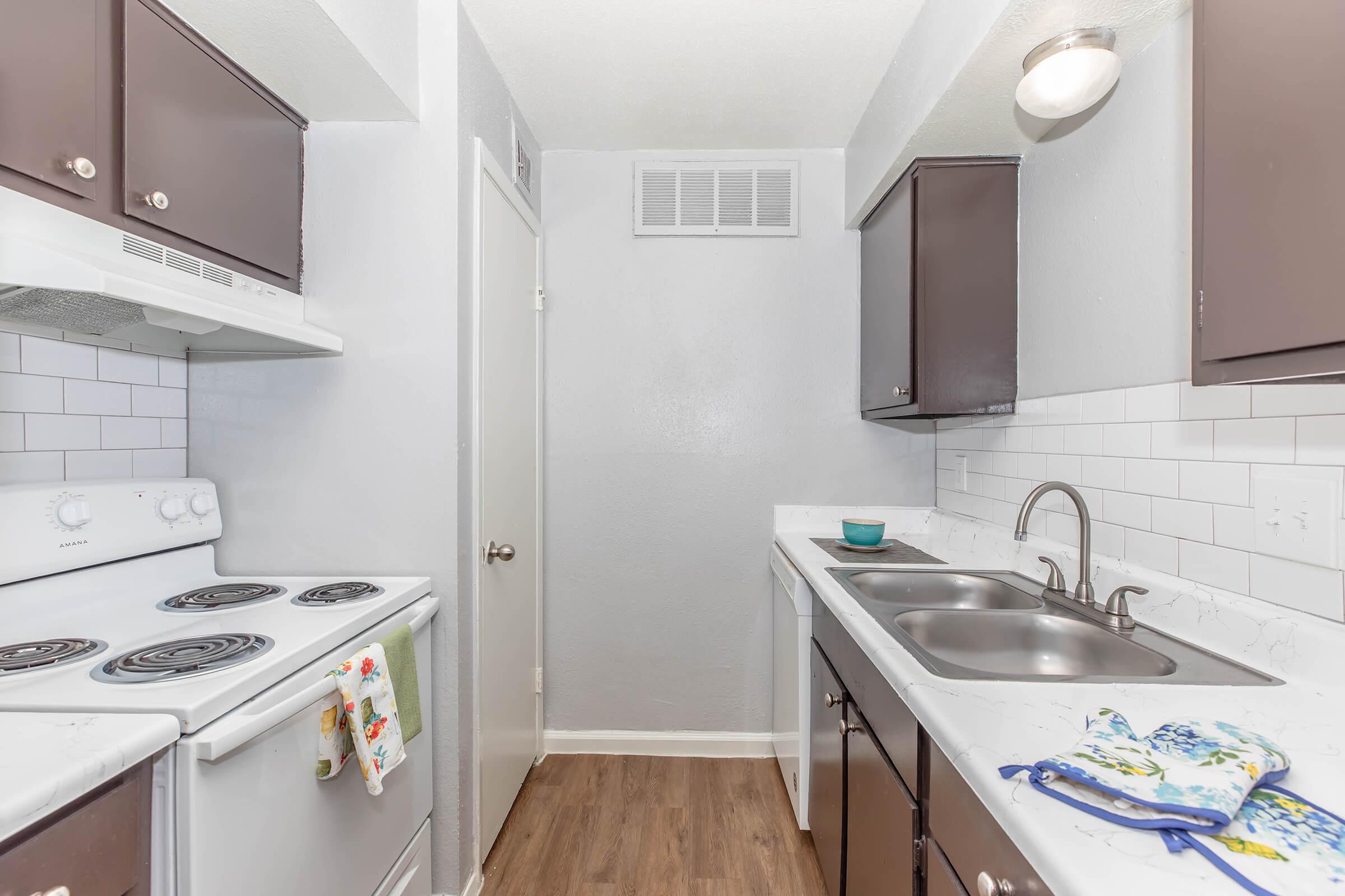 A modern kitchen featuring white appliances, including an electric stove and a double sink. The cabinetry is dark brown, complemented by white subway tiles on the backsplash. Natural light flows in, illuminating the wood-look flooring and creating a clean, inviting atmosphere.