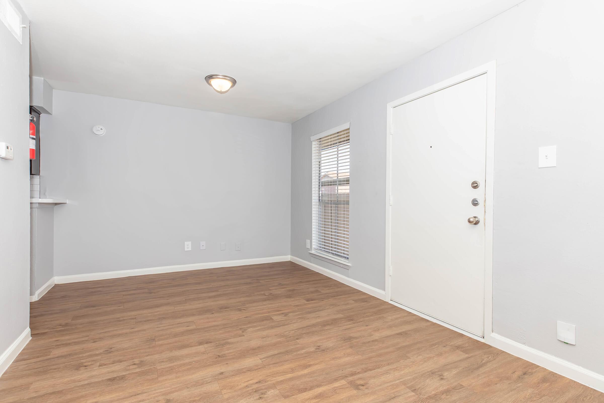 An empty room featuring light gray walls, a wooden floor, and a ceiling light fixture. A door with multiple locks is visible on the right, while a window with blinds offers natural light. The adjacent wall shows a small kitchen area, partially visible on the left.