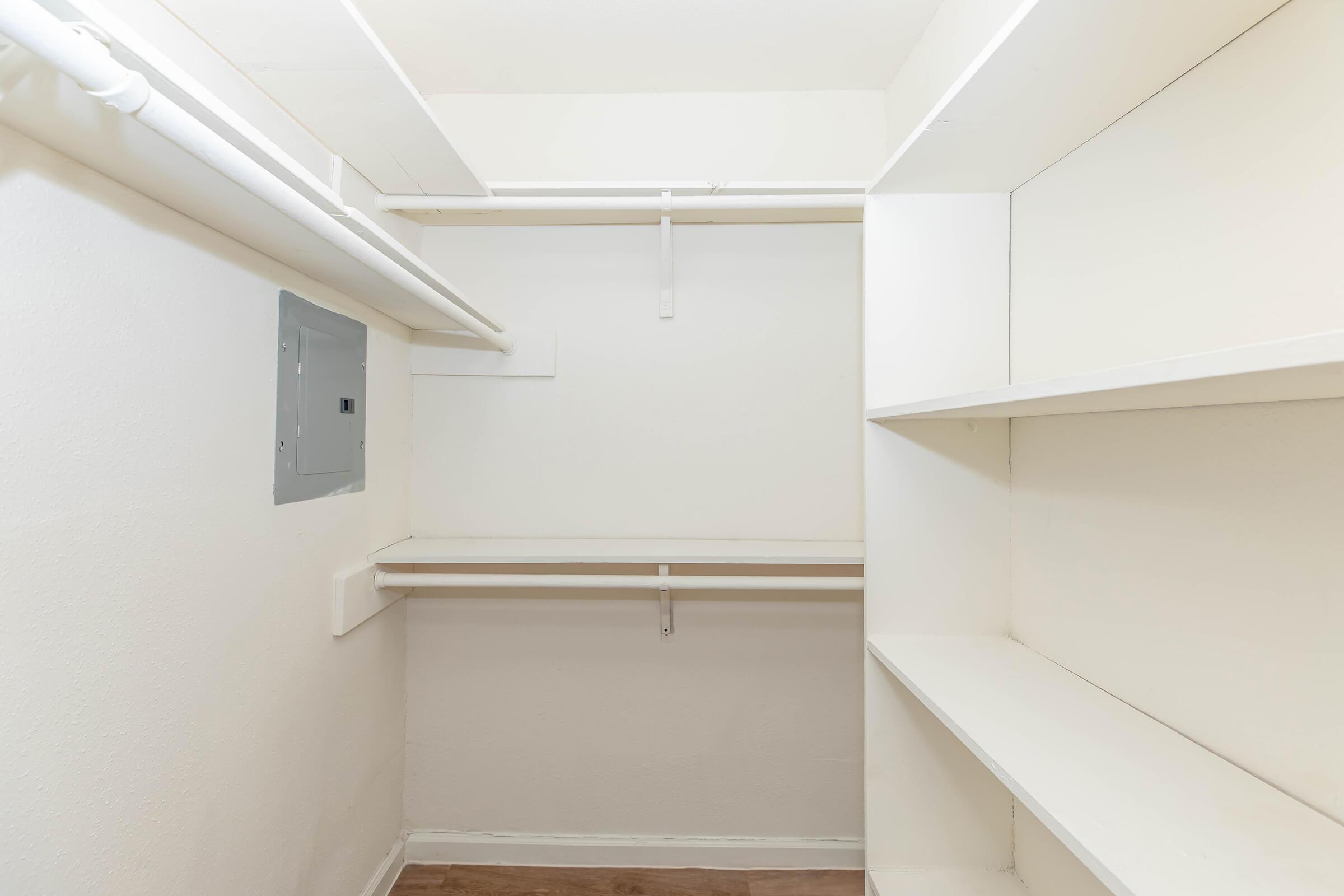 Empty closet with white walls, features two shelves on the right side, a hanging rod along the left wall, and an electrical panel on the far side. The floor is light-colored, giving a clean and spacious appearance.