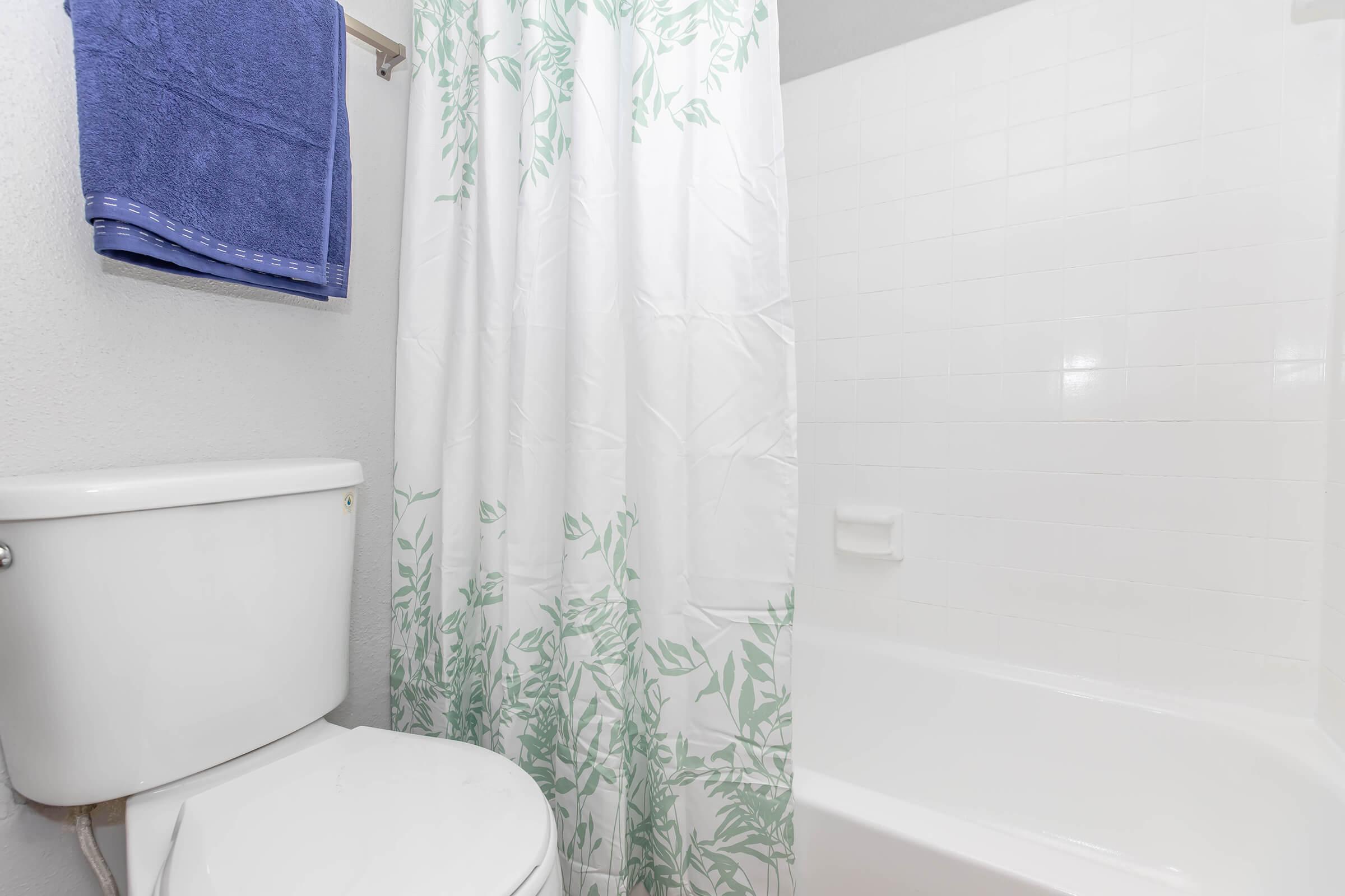 A clean bathroom featuring a white toilet next to a bathtub with a white tiled wall. A shower curtain with green leaf patterns is present, along with a blue towel hanging on a rack. The overall atmosphere is light and tidy, emphasizing a fresh and inviting space.