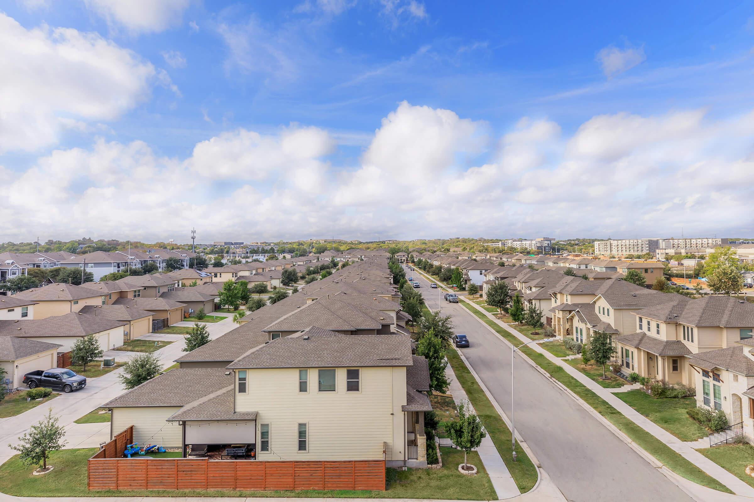 Aerial view of a suburban neighborhood featuring rows of houses with varying designs and colors, green lawns, and tree-lined streets under a partly cloudy blue sky. The layout shows a mix of single-family homes and community infrastructure in a well-maintained area.