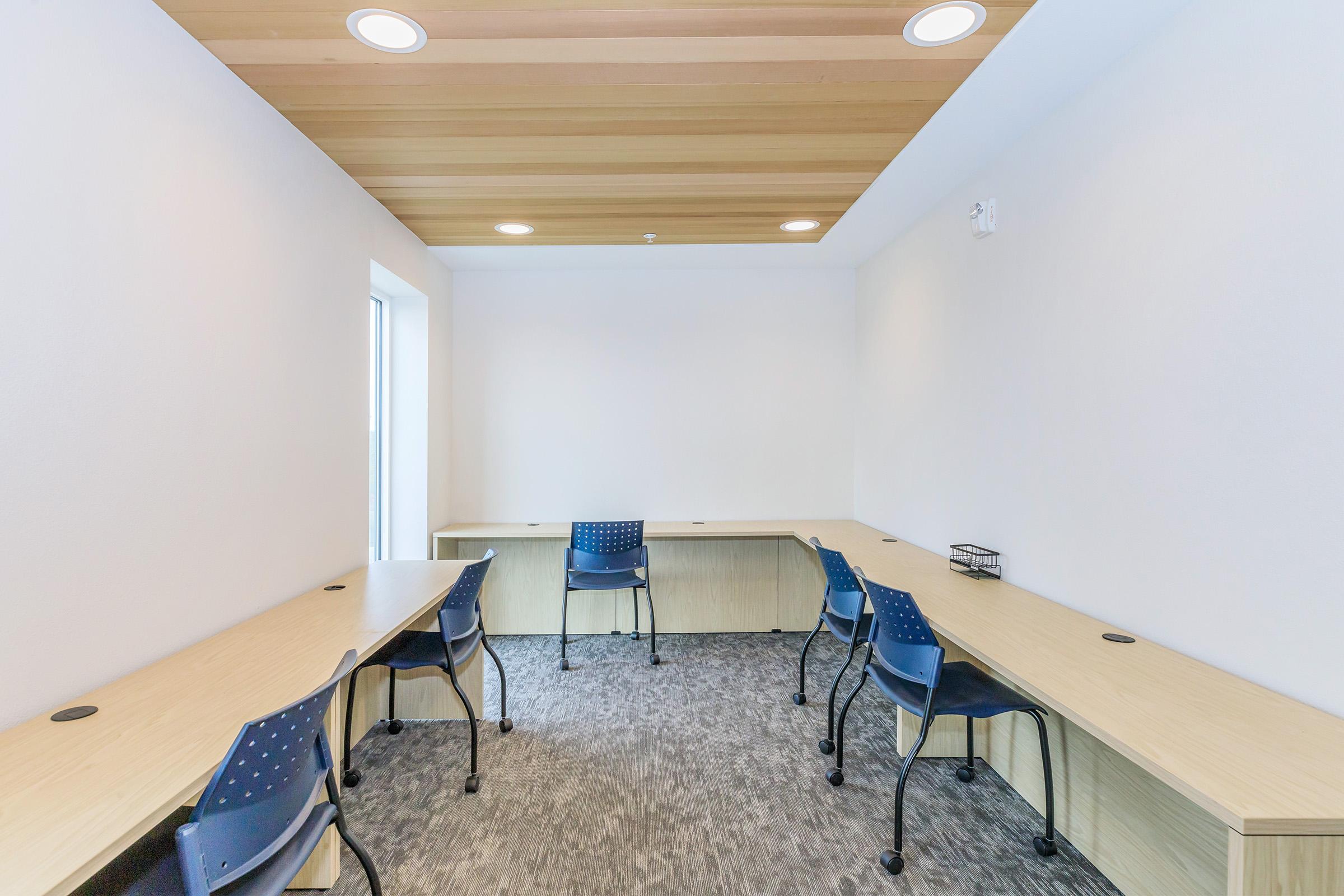 A modern, minimalist office space featuring a light wood ceiling, two desks with blue chairs, and a neutral color scheme. The room is well-lit with overhead lights and has a single window allowing natural light. The floor is covered with grey carpeting, providing a clean and professional environment.