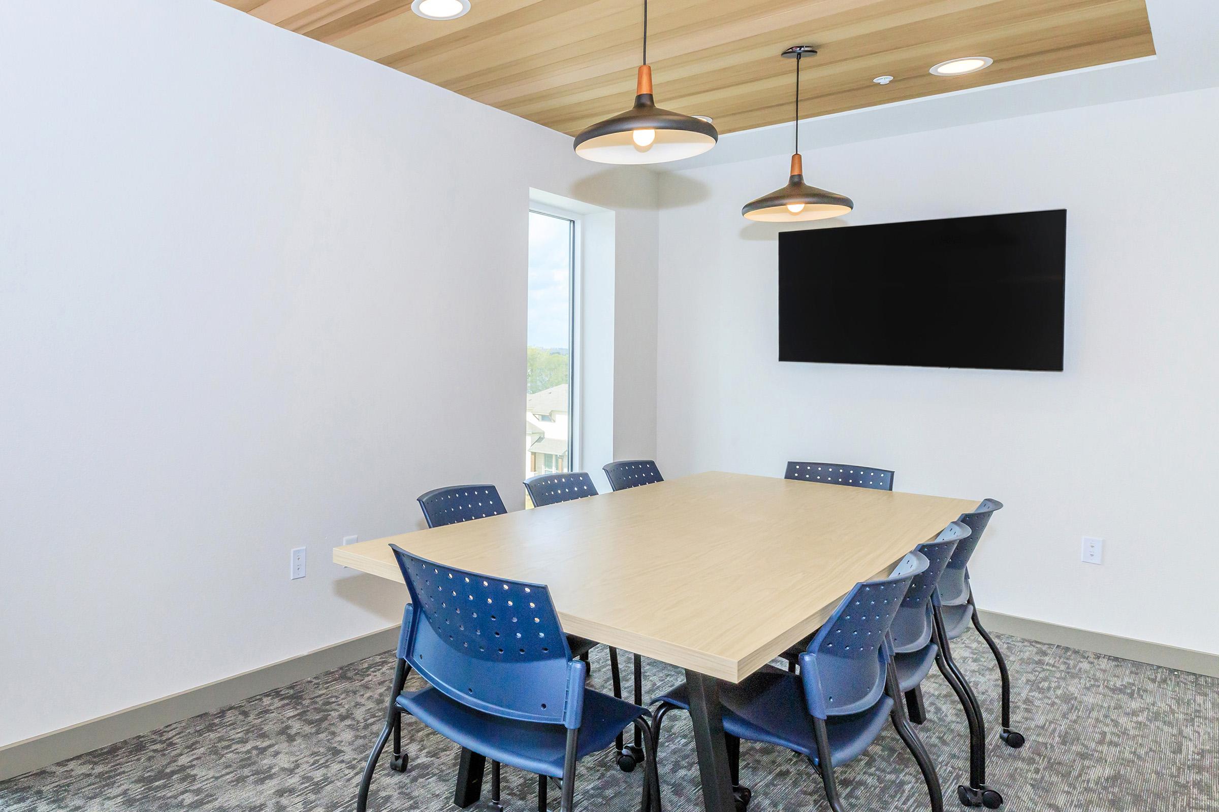 A bright conference room featuring a large wooden table surrounded by six blue chairs. The room has a window allowing natural light in and a flat-screen TV mounted on the wall. The ceiling has modern light fixtures, and the floor is covered in carpet.