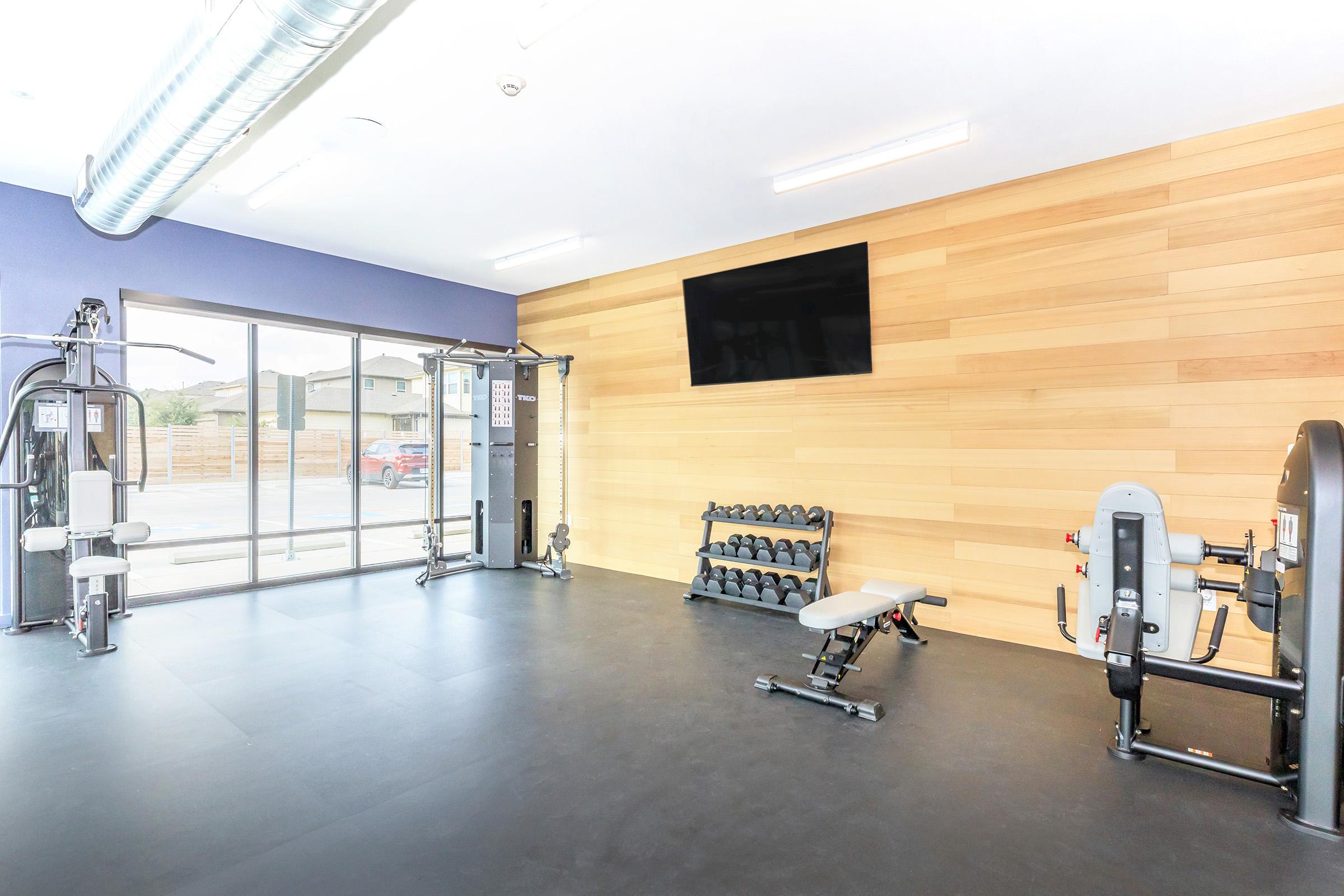 A modern gym interior featuring exercise equipment, including weight machines, dumbbells on a rack, and a weight bench. Large windows allow natural light, and a wall-mounted TV adds to the ambiance. The space has wooden paneling and a clean, organized layout, suitable for workouts.