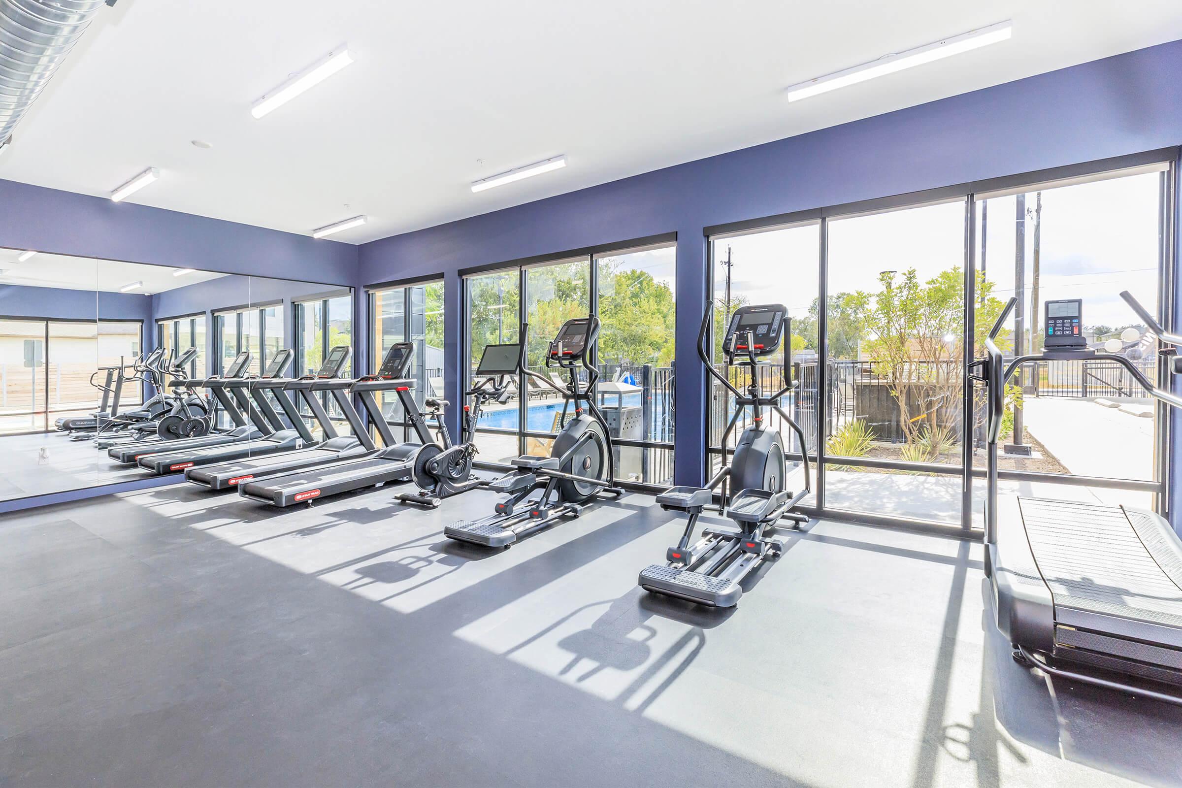 A modern gym featuring several pieces of cardio equipment, including treadmills and elliptical machines, arranged against large windows that allow natural light. The interior has a clean and open feel, with a view of outdoor greenery.
