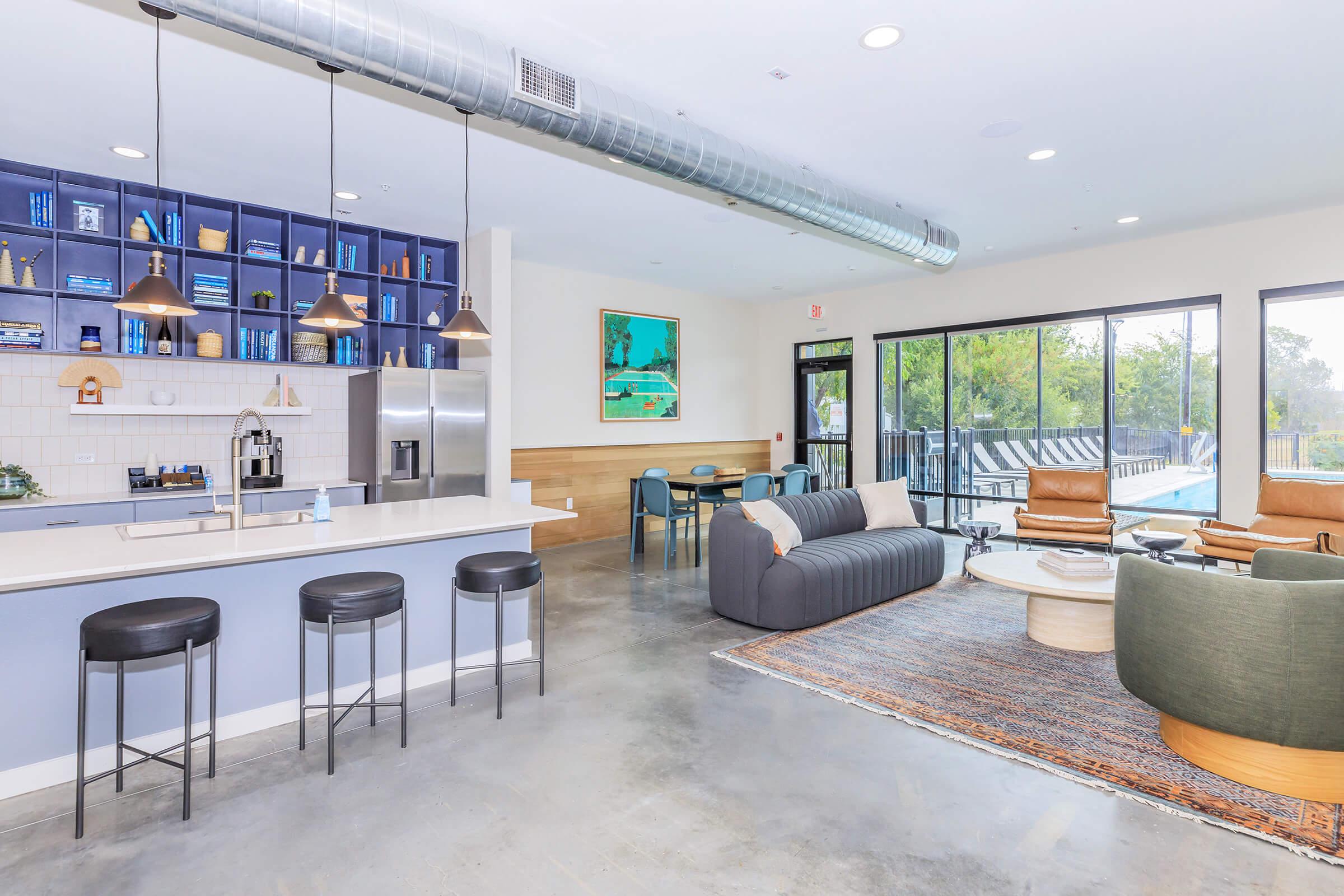 Modern communal living space featuring a kitchen island with bar stools, bookshelves, and a cozy seating area. Large windows provide natural light and a view of an outdoor pool area. Stylish decor includes artwork and a rug, creating a welcoming atmosphere for social gatherings.
