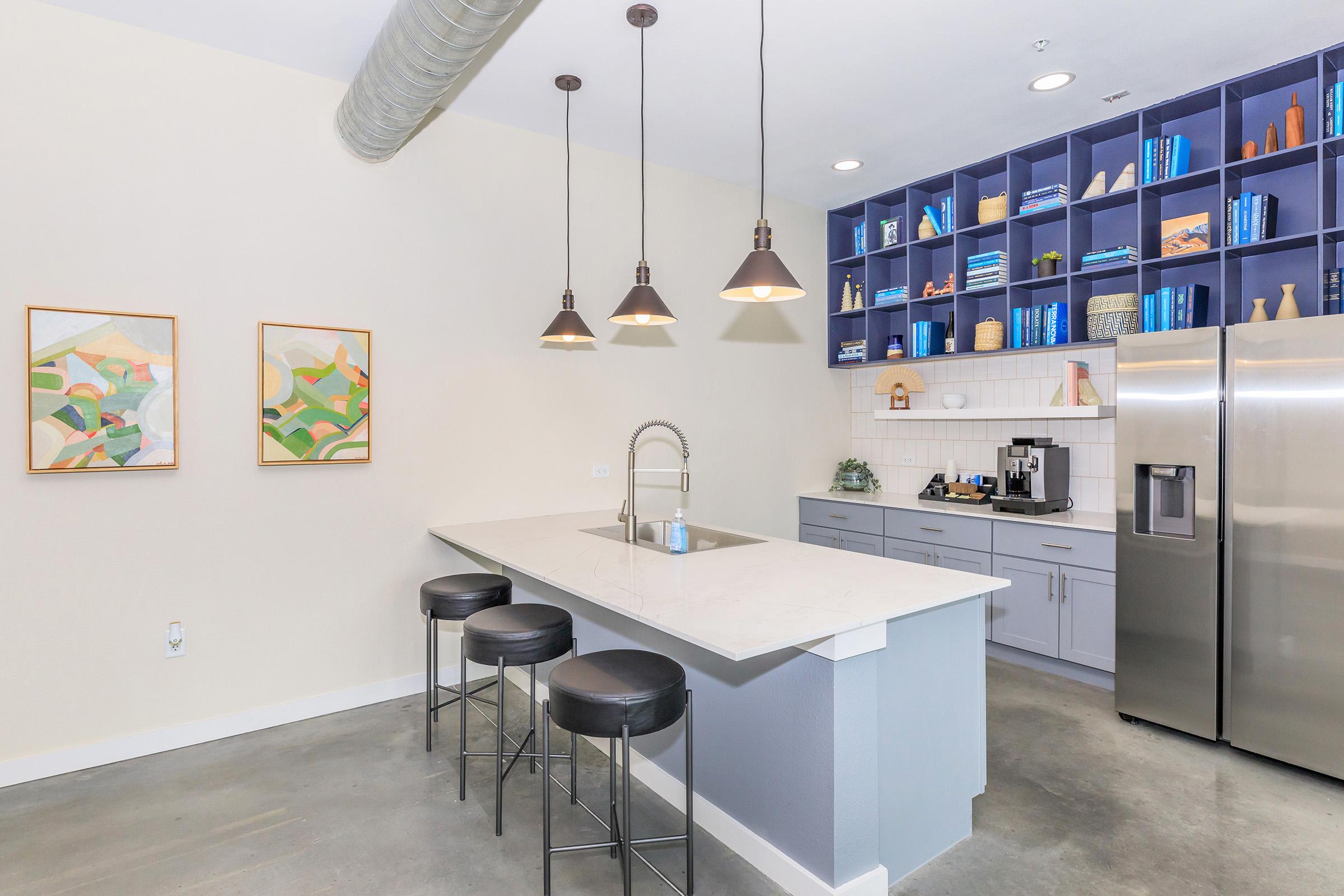 Modern kitchen with a minimalist design featuring a large island with three black bar stools. Above the island, three pendant lights hang down. A stainless steel refrigerator is visible, along with cabinetry filled with blue books and decorative items. Light-colored walls and concrete flooring complete the contemporary look.